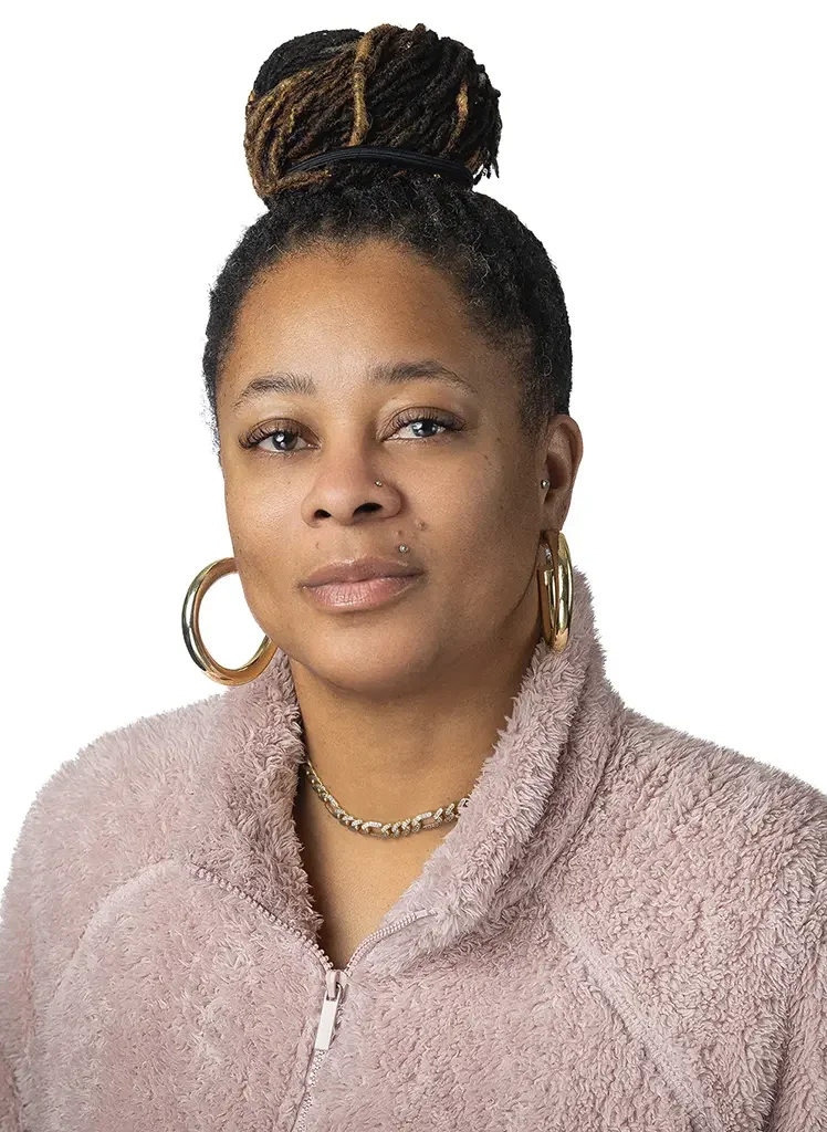 A woman with a high bun of dreadlocks, wearing large gold hoop earrings, a gold chain necklace, a nose ring, and a pink fuzzy zip-up jacket, looking at the camera against a white background.