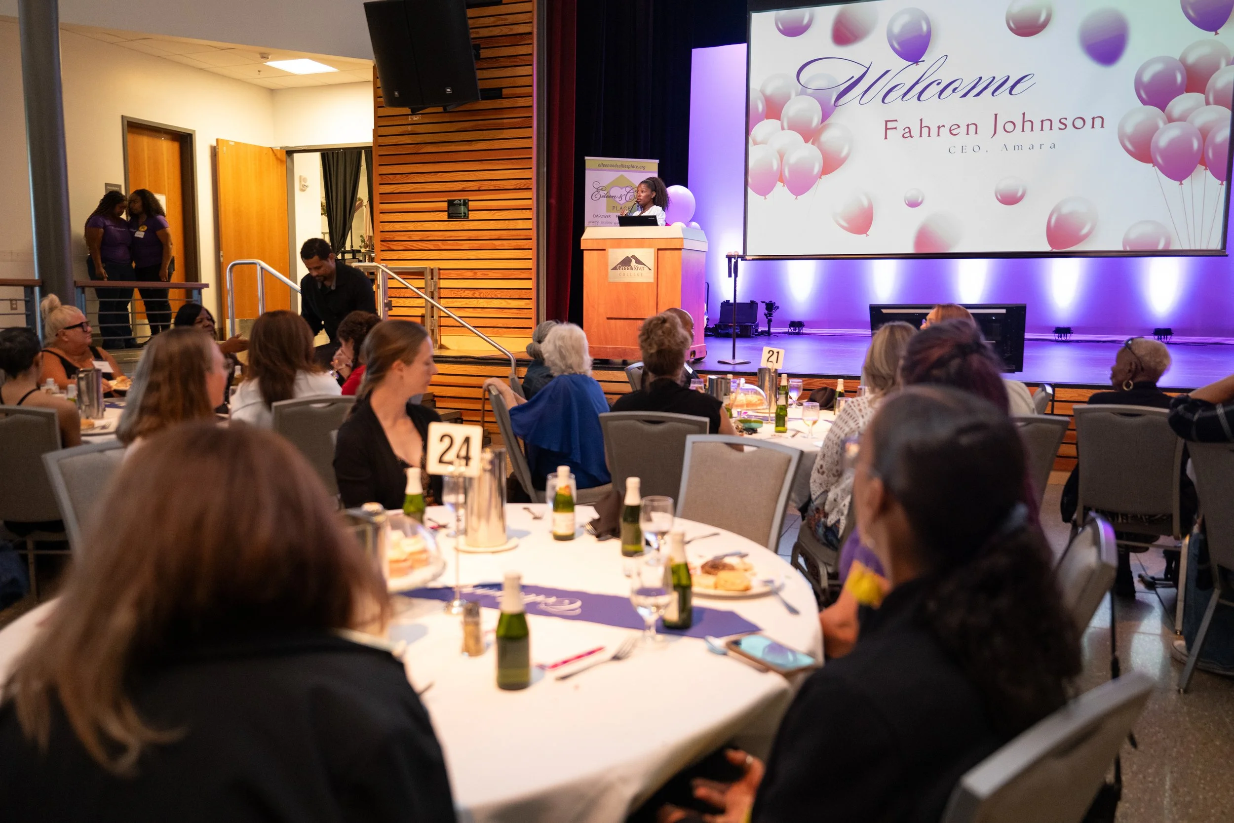 Conference attendees seated at round tables in a banquet hall facing a stage with a large screen displaying a welcome message for Fahrend Johnson. There is a woman speaking at a podium on the stage, and the room is decorated with balloons and soft lighting.