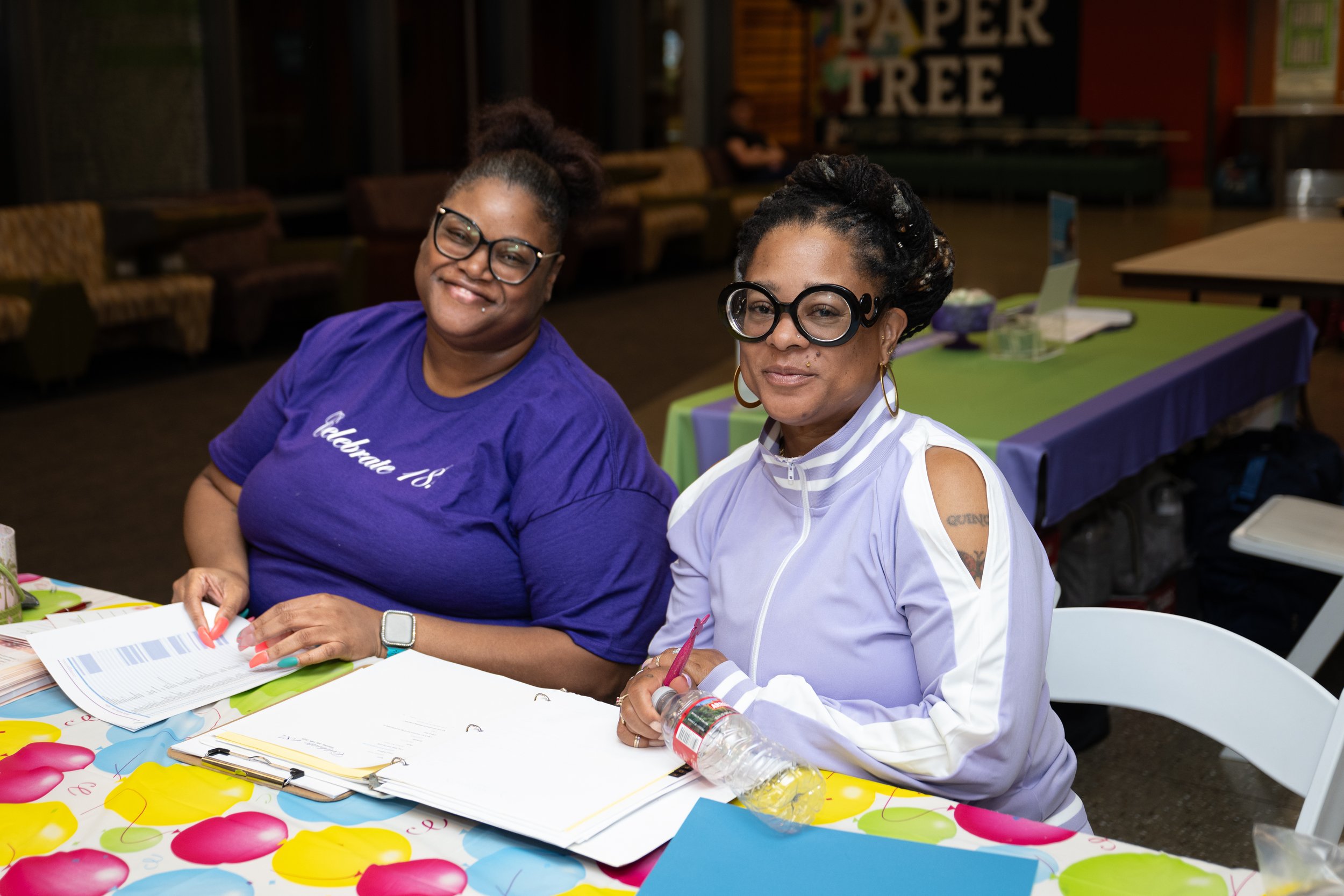 Two women sitting at a colorful table with papers, notebooks, and a water bottle, smiling and looking at the camera inside a decorated event space.