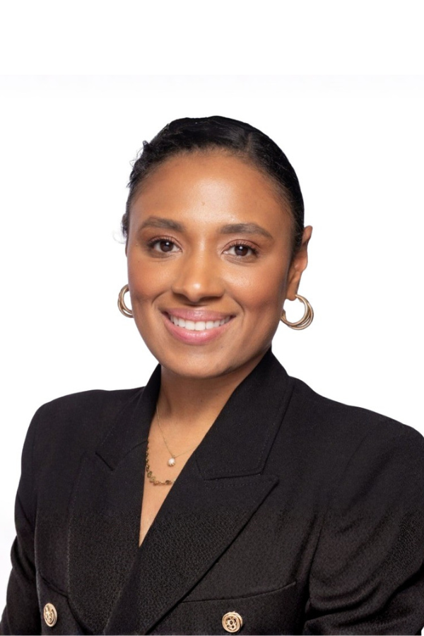 A woman with dark hair pulled back, wearing gold hoop earrings, a pearl necklace, and a black blazer, smiling against a white background.