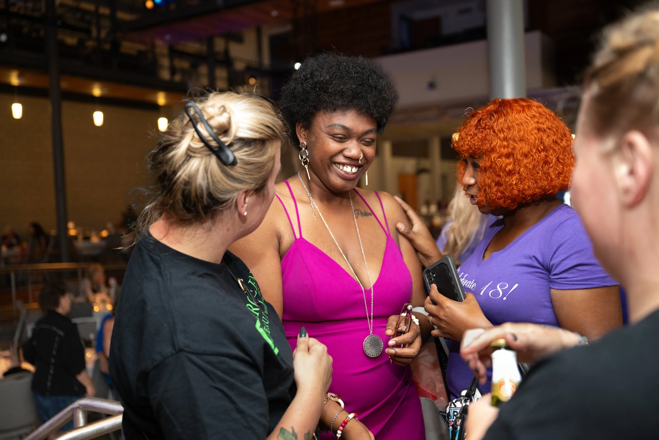 Group of women celebrating together, one wearing a pink dress and others in casual attire, in a lively indoor setting.