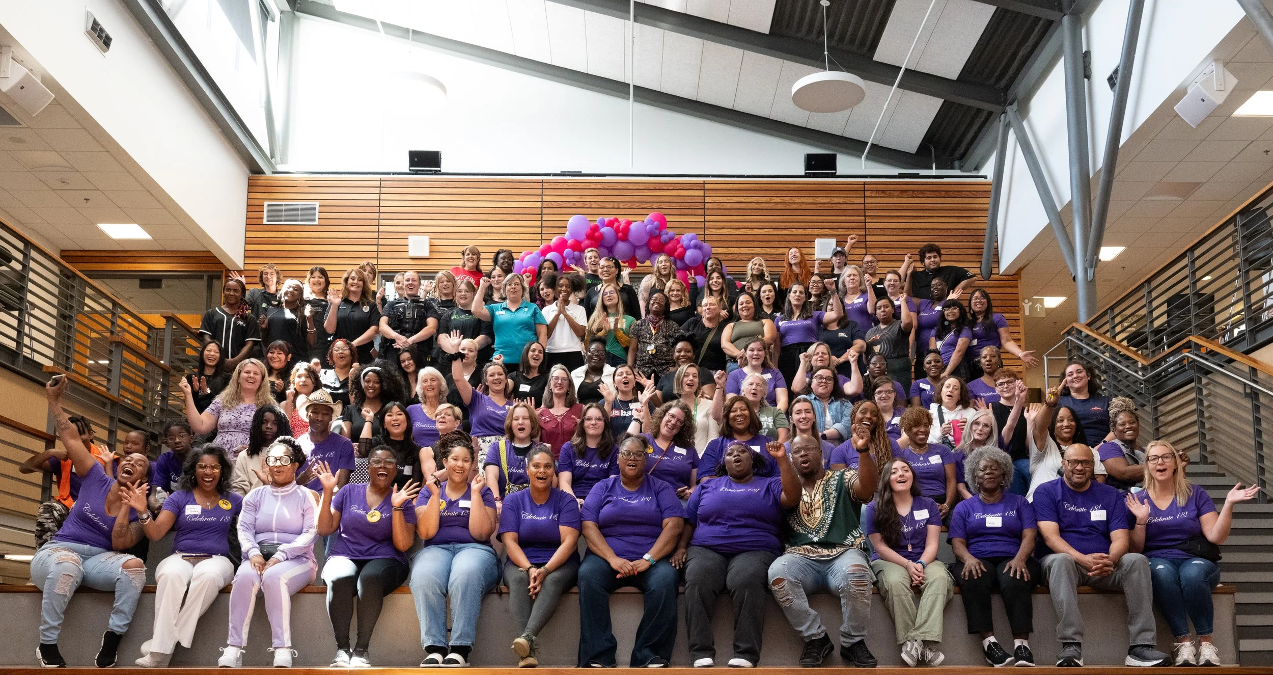 Large group of diverse people gathered on steps inside a modern building, celebrating at an event, with pink and purple balloons in the background.