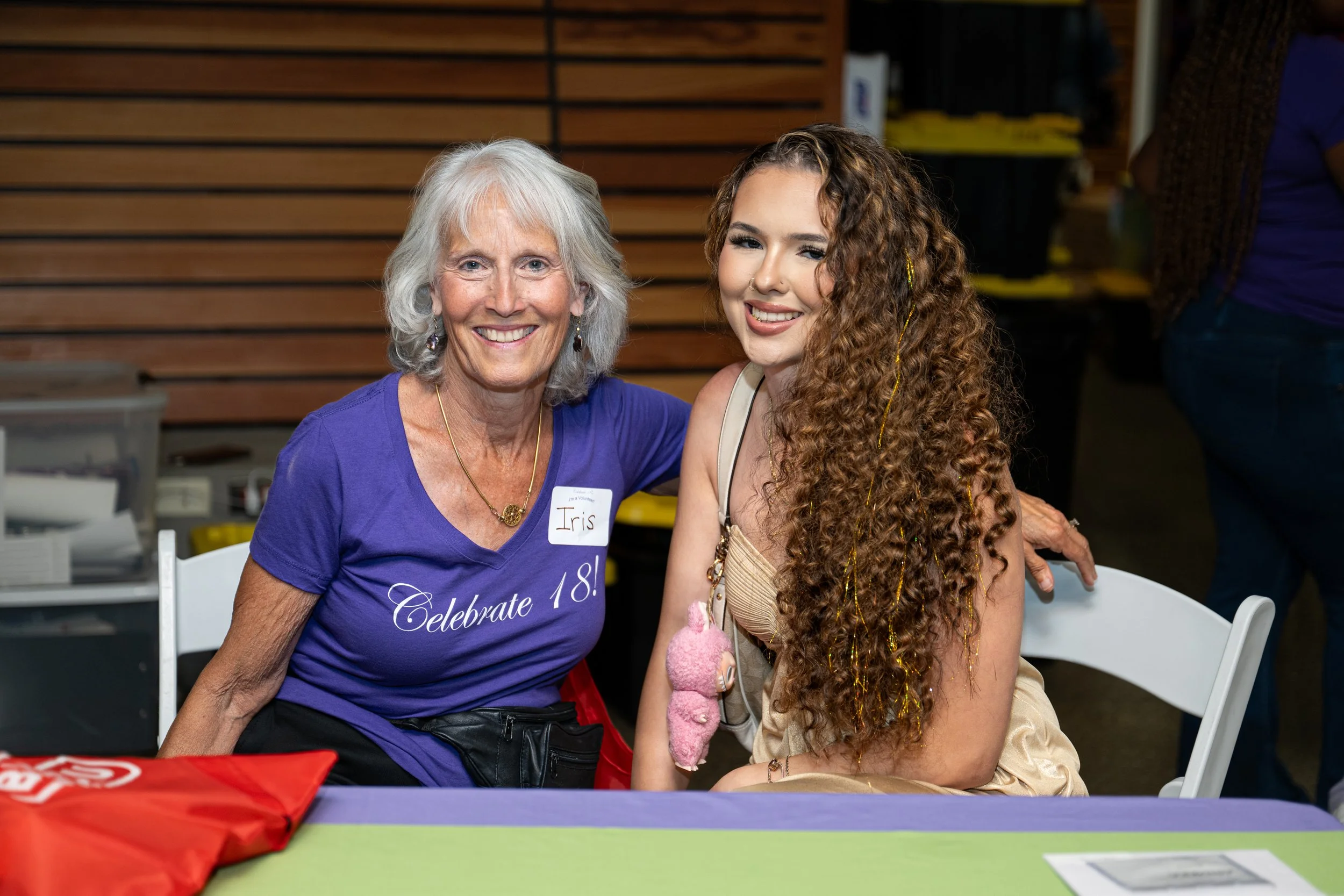 An older woman and a young woman sitting at a table, smiling at the camera, with a wooden wall background.