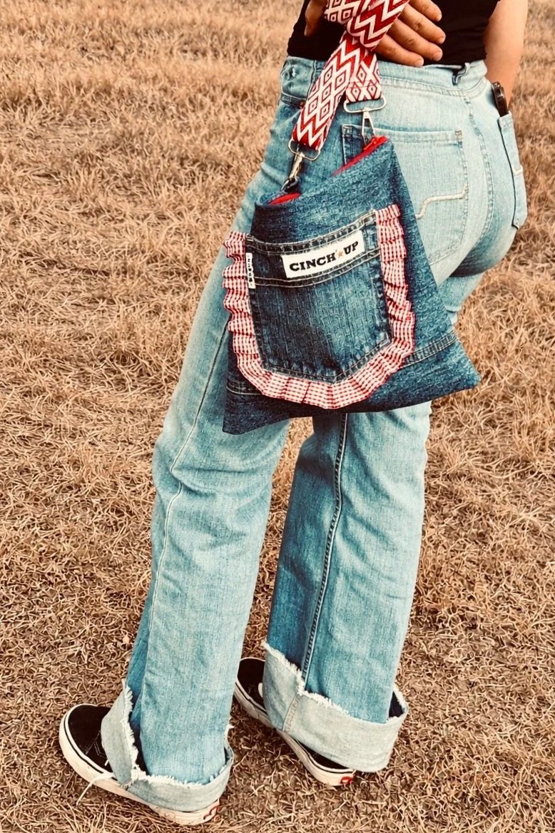A person standing outdoors on dry grass, wearing western jeans with cuffs, black sneakers, and holding an up-cycled denimdarl bag with red check pocket frill and red and white check bag strap.