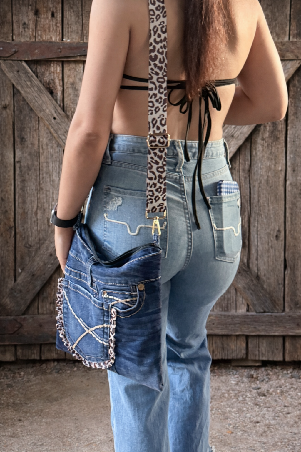 Back view of a woman wearing a black top, light blue western jeans, and an up-cycled denimdarl crossbody denim bag with leopard print strap hanging over her shoulder, standing in front of a wooden barn door.
