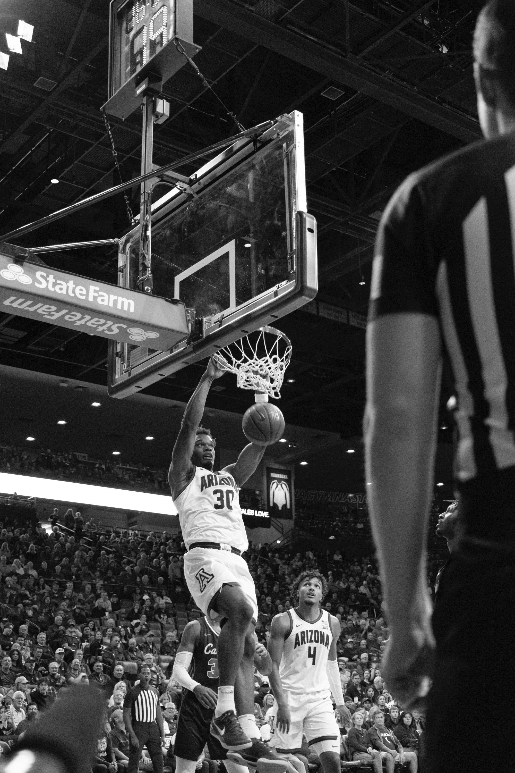 A basketball player in a white Arizona jersey with the number 30 is dunking a basketball during a game, with other players and spectators in the background.