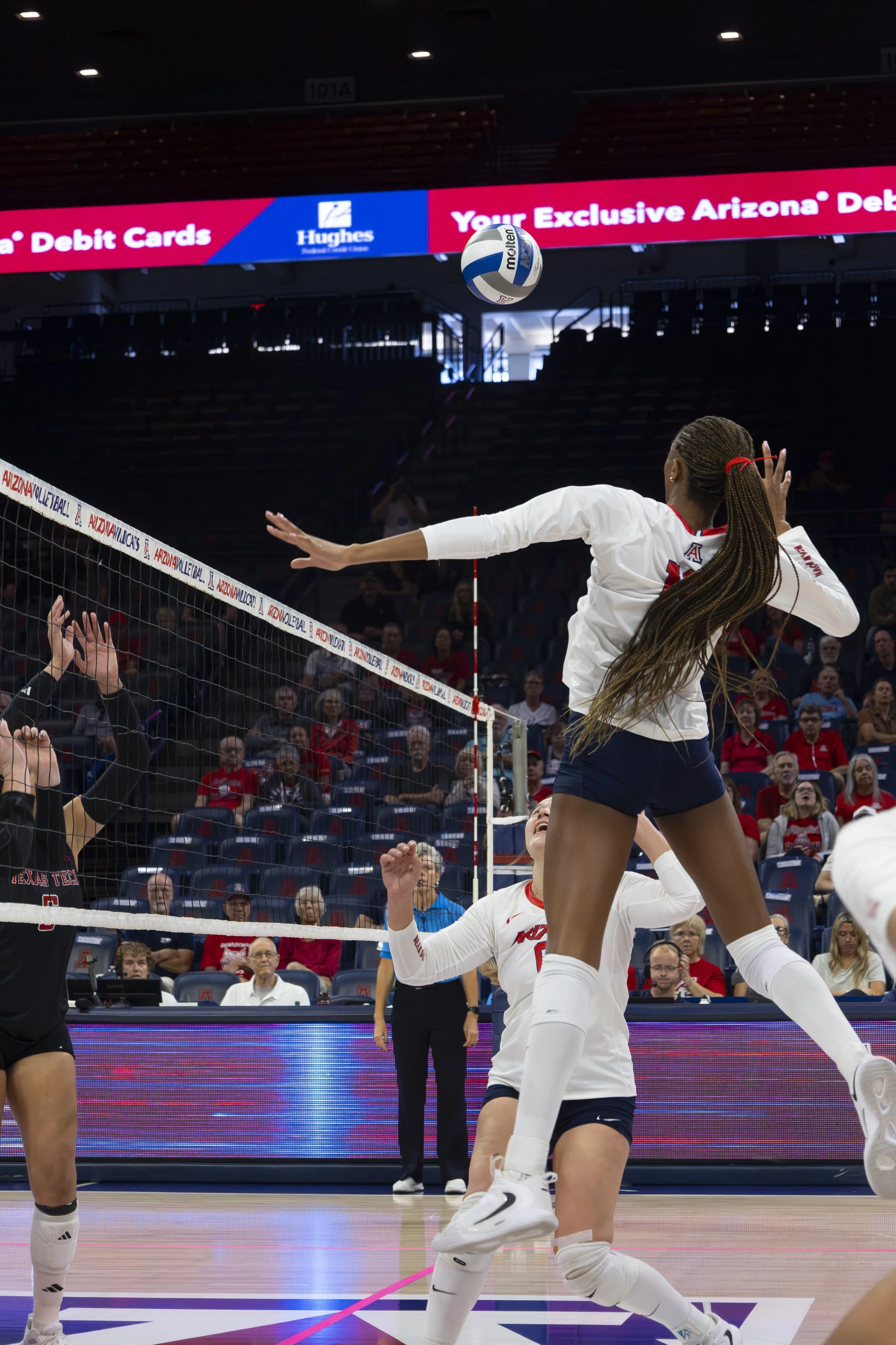 A volleyball player in a white jersey jumps to hit the ball over the net during a match in an indoor arena, with spectators watching in the background.