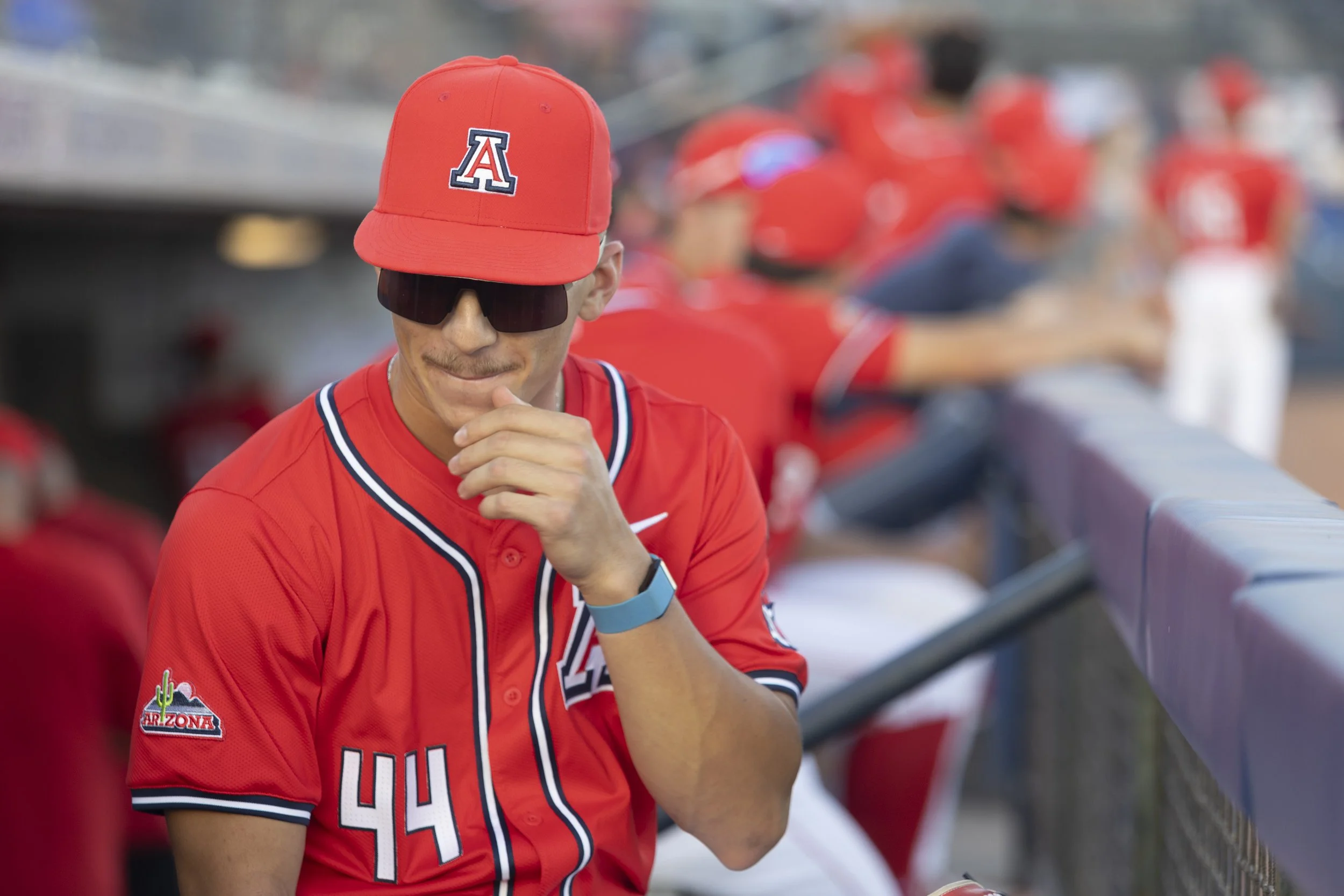 A baseball coach or player dressed in a red Arizona Diamondbacks jersey and cap, standing near the dugout with other team members in the background.