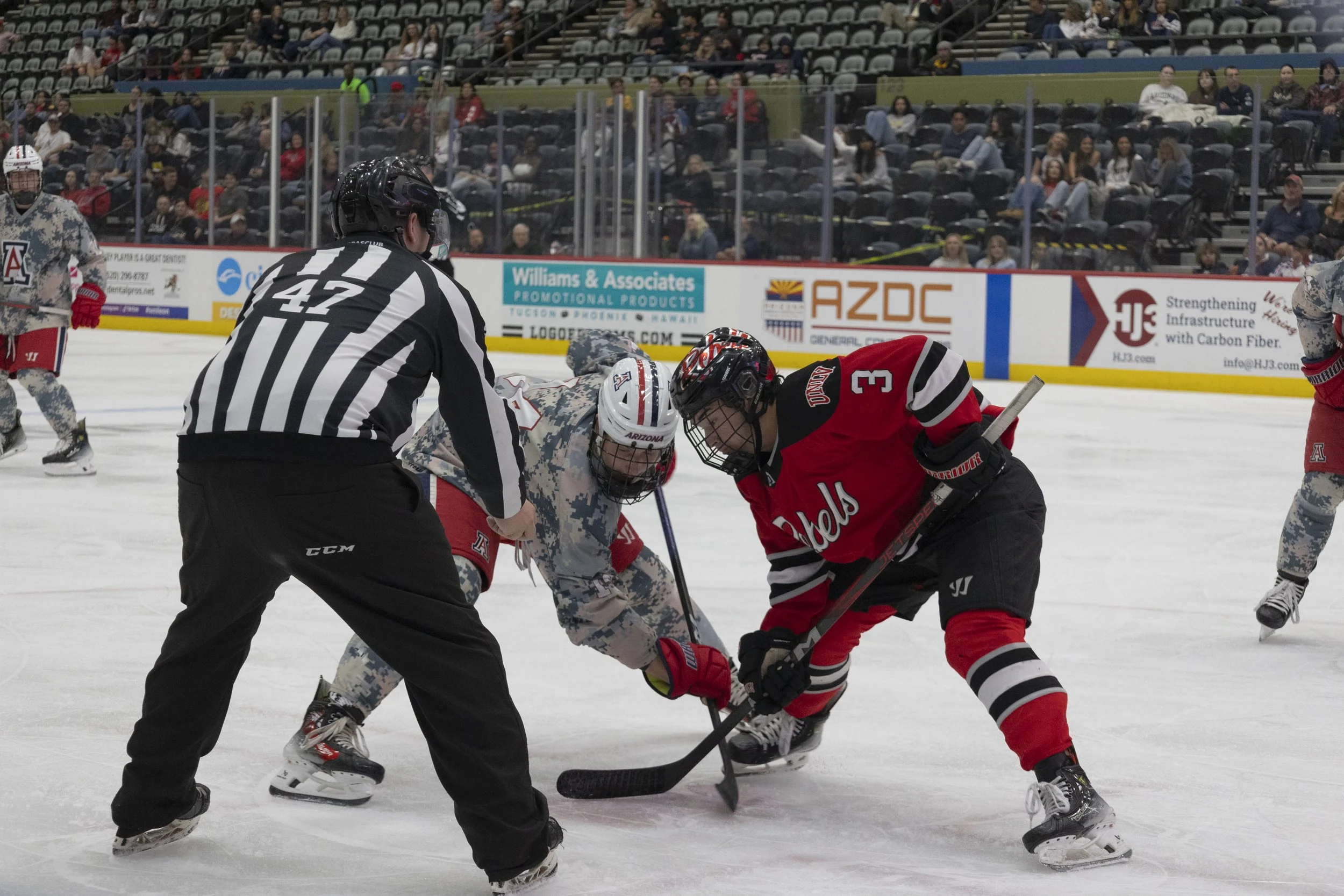 Hockey game with players in red and camouflage jerseys battling for puck, referee in black and white stripes watching, spectators in stands.