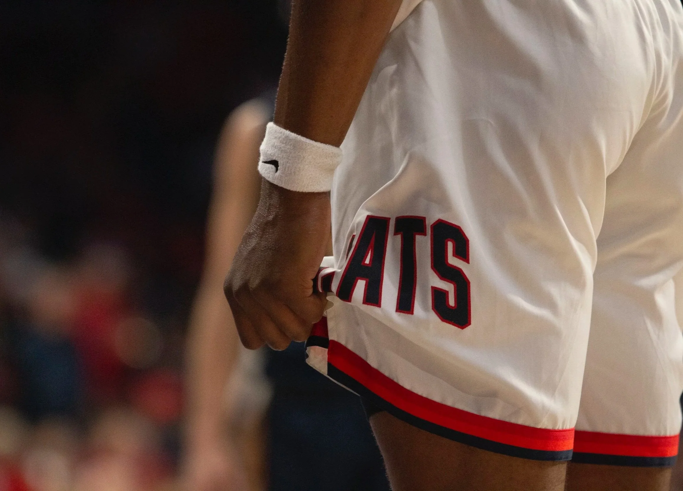 Close-up of a tennis player wearing a white wristband and white shorts with the word 'GAT'S' visible on the side. The player's hand is clenched into a fist near the shorts, with a blurred background.