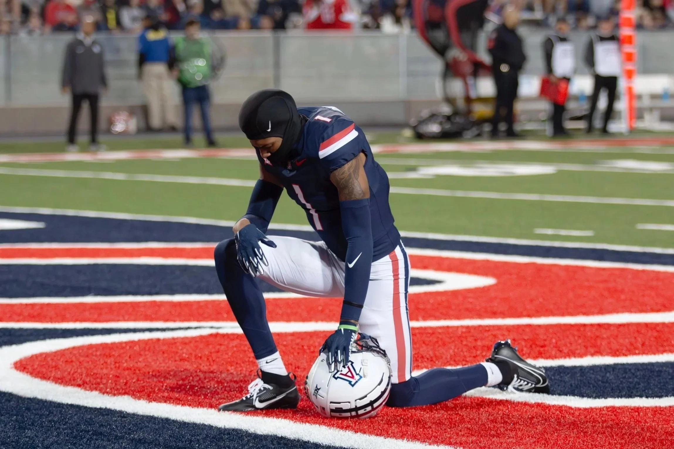 An American football player kneeling on the field with his helmet on the ground, wearing a navy blue uniform with red and white accents, during a game.