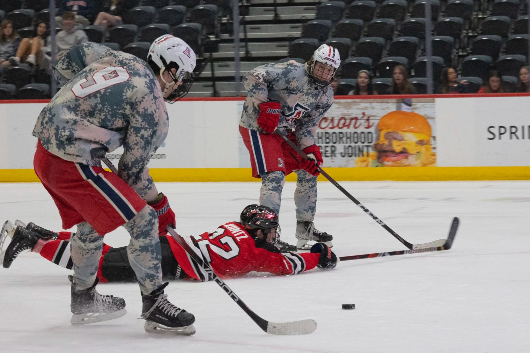 Hockey players in camouflage uniforms and red jerseys engaged in a game, with one player on the ice and others competing for the puck, in an arena with spectators in the background.