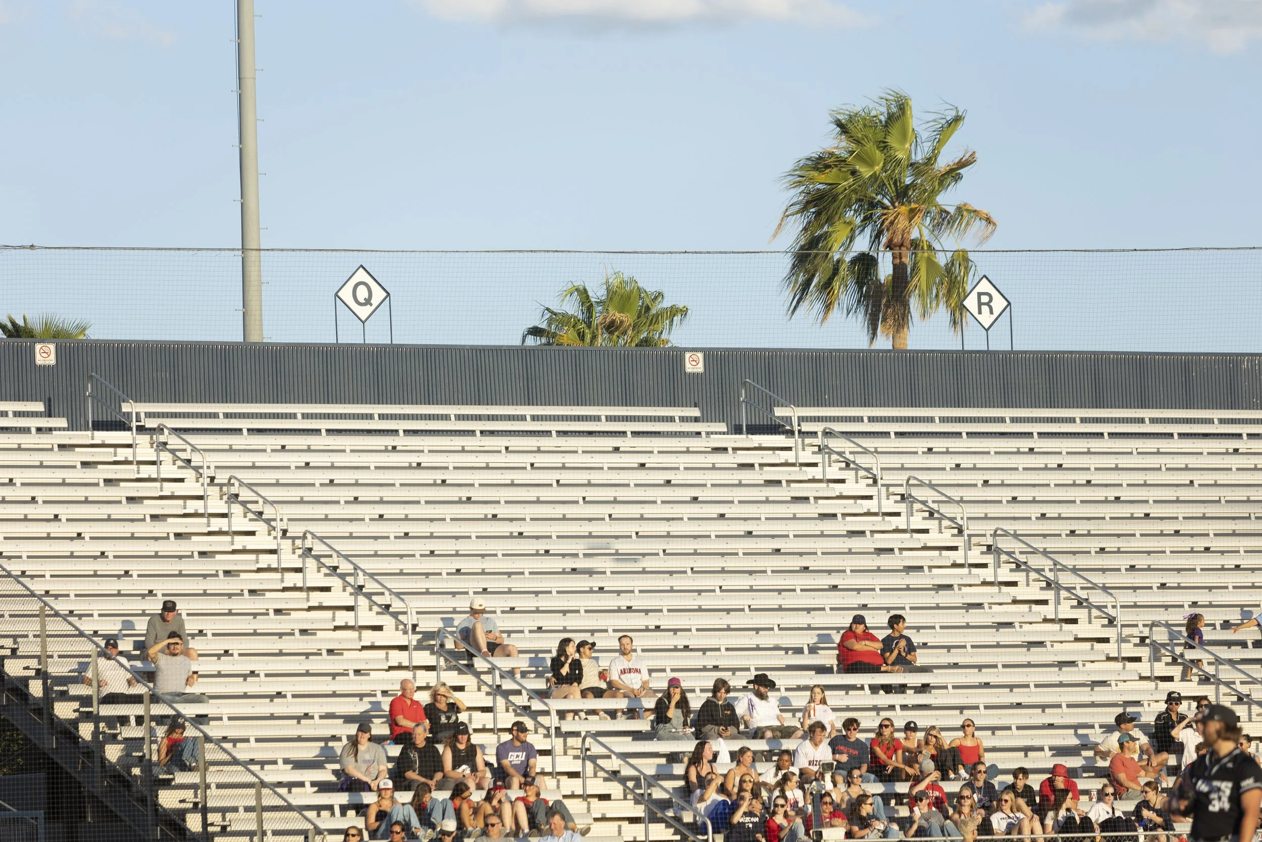 A mostly empty bleacher seating area with a few people sitting, sunlight, palm trees in the background, and signs with letters Q and R on the back fence.