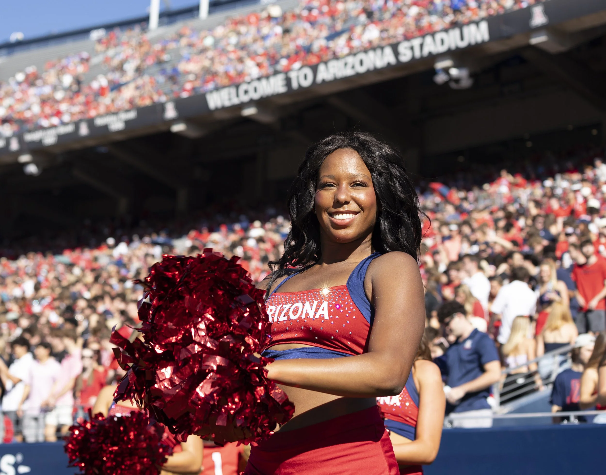 A cheerleader in a red and blue Arizona Wildcats uniform holding red pom-poms at a football game, smiling with a crowd in the stadium background