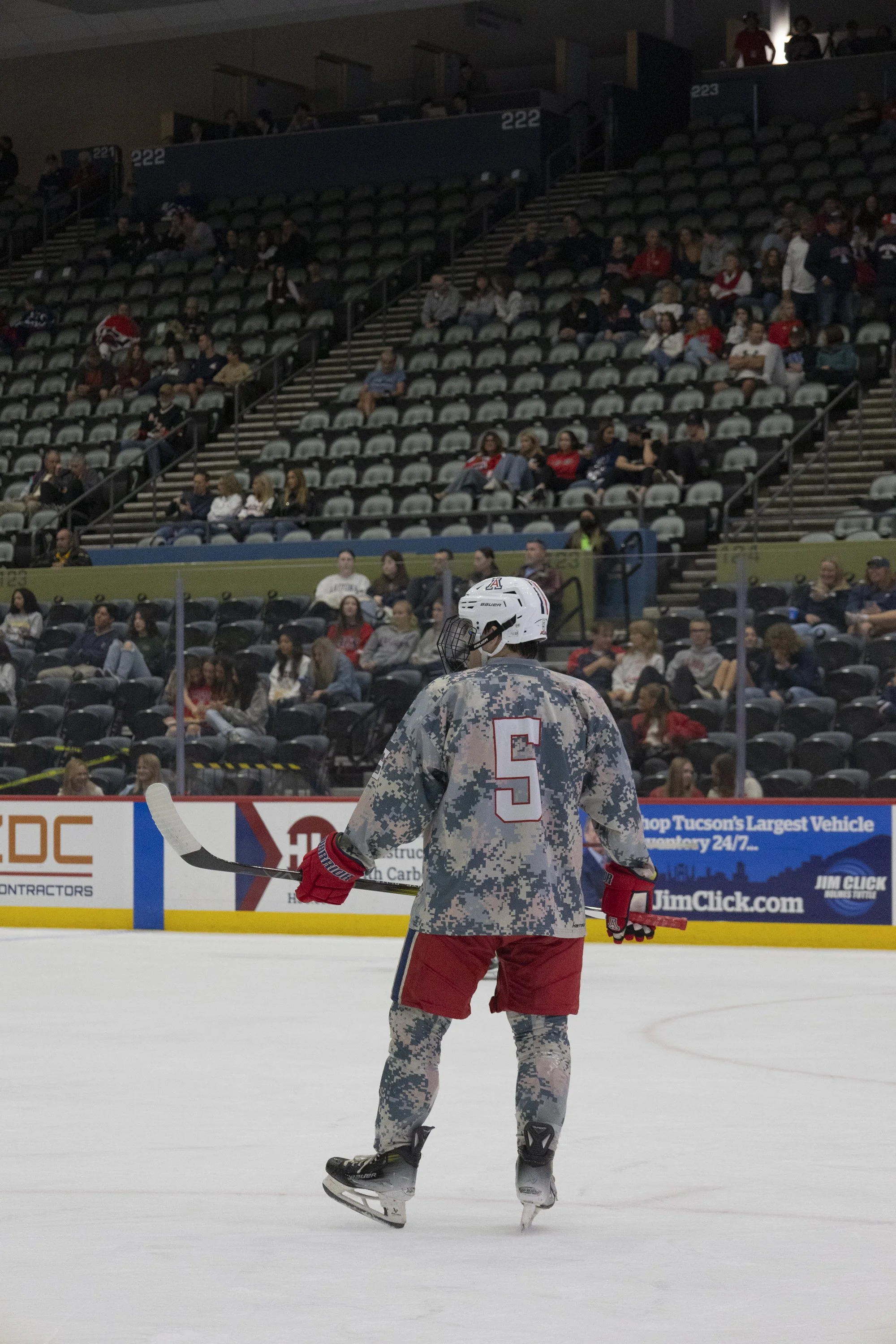 A hockey player in camouflage jersey with number 5 standing on ice rink with empty seats and spectators in the background.