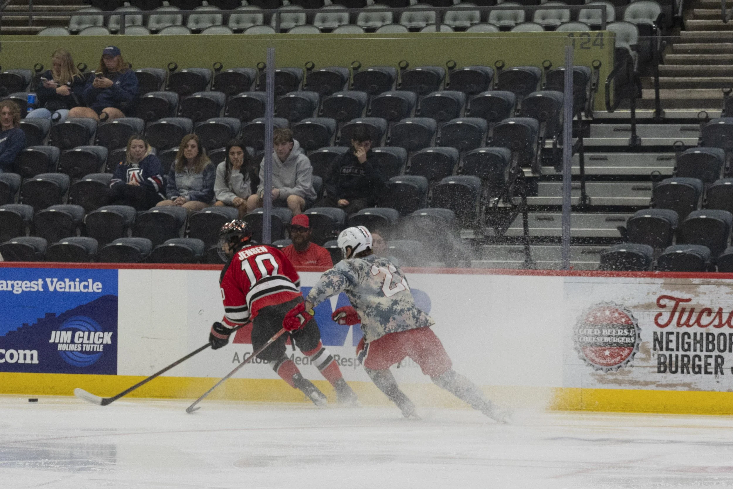 Two hockey players battling for the puck near the boards, one in a red jersey and the other in a camouflage pattern jersey, with spectators watching from the stands.