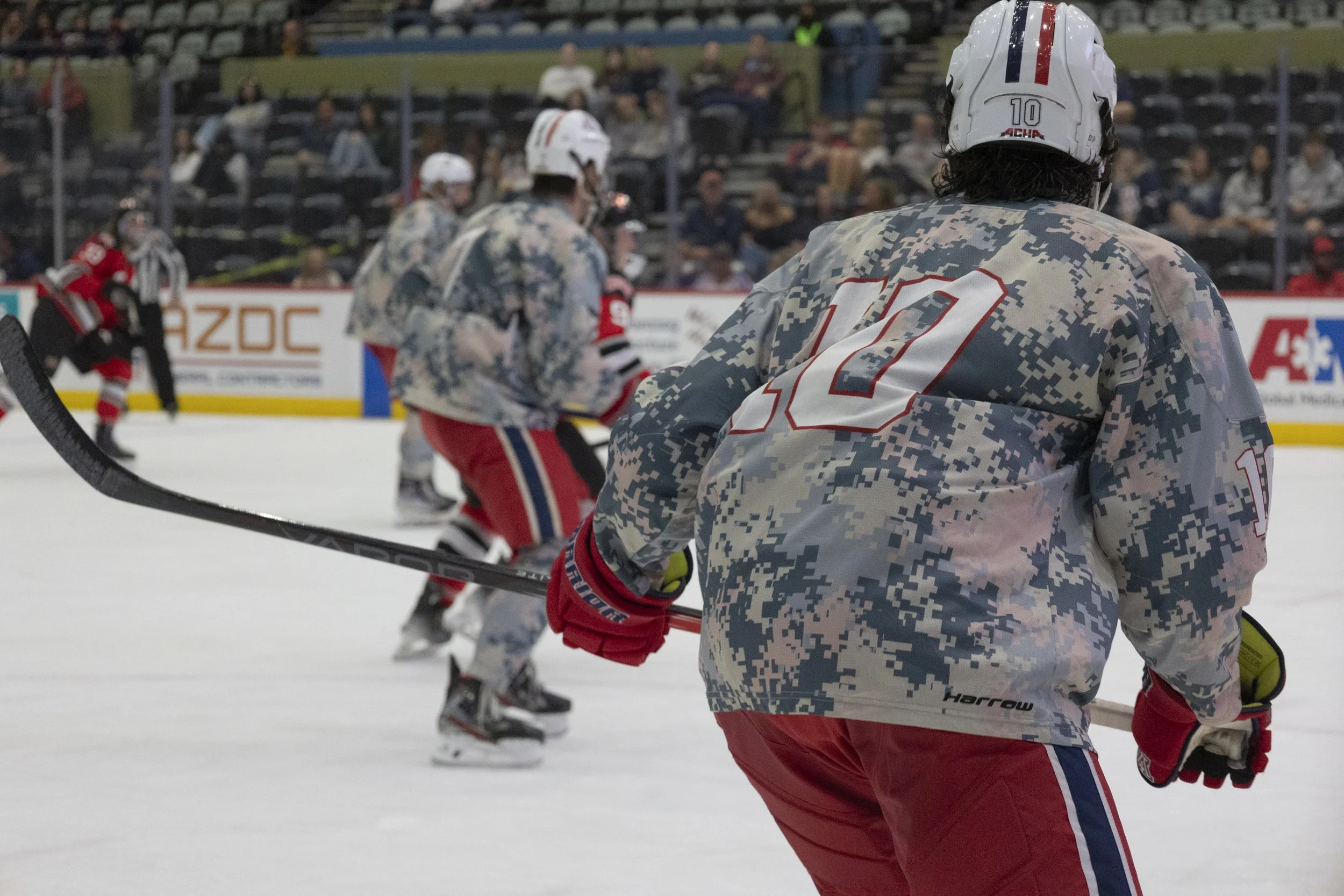 Ice hockey players in camouflage jerseys on the ice rink during a game, with spectators in the background.