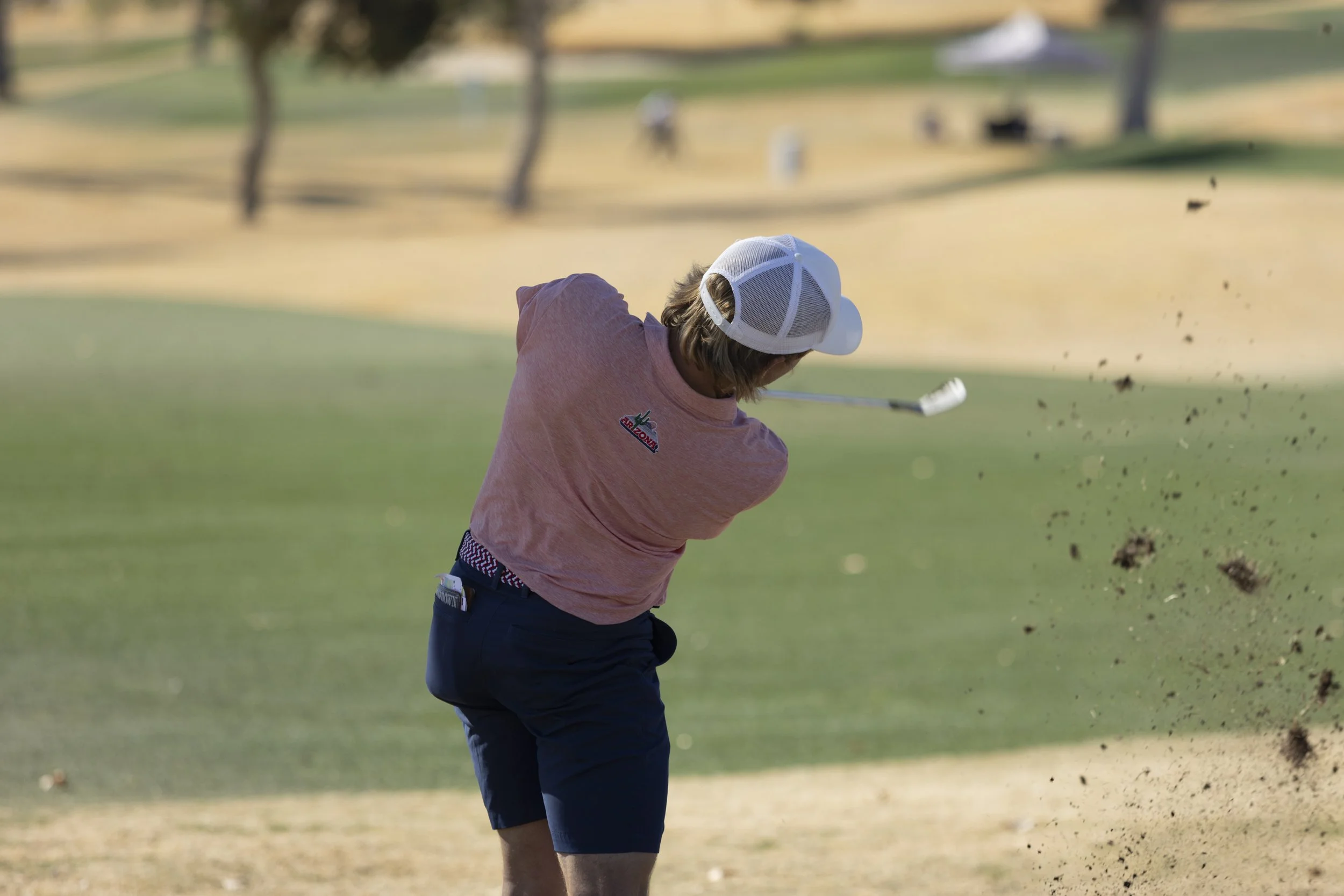 Young man playing golf on a sunny golf course, swinging a club and hitting the ball into a sand trap, wearing a pink shirt, dark shorts, and a white cap.