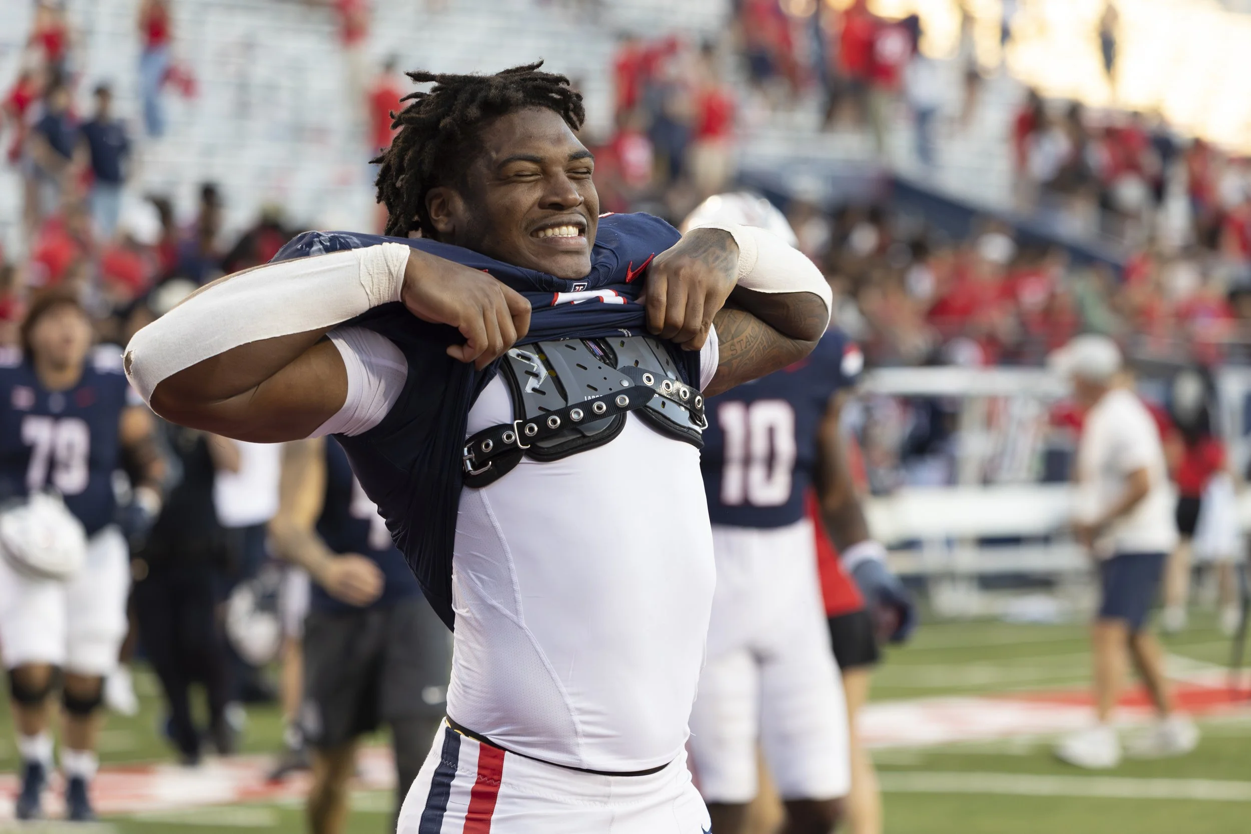 A male football player with dreadlocks, wearing a white jersey with red, white, and blue stripes and a black harness, is pulling his jersey over his head in a stadium with fans in the background.