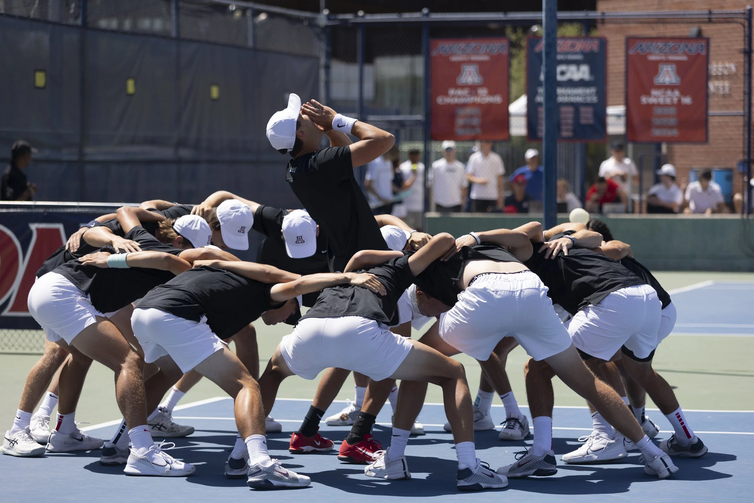 Tennis team huddled together on a court, with one player standing and making a gesture, US and Arizona banners in background.