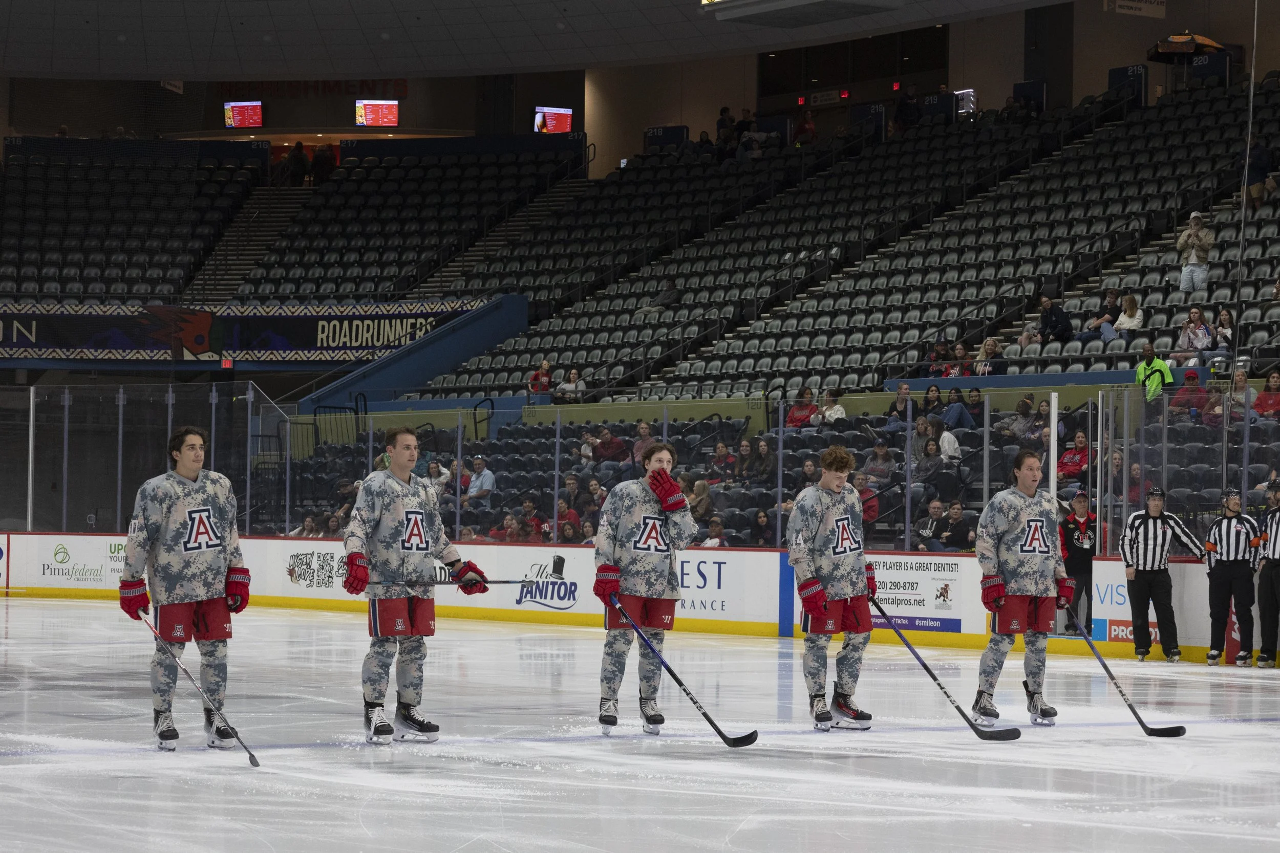 Hockey players in camouflage jerseys and red shorts standing on ice rink, lining up before game in an indoor arena, with spectators in the stands.