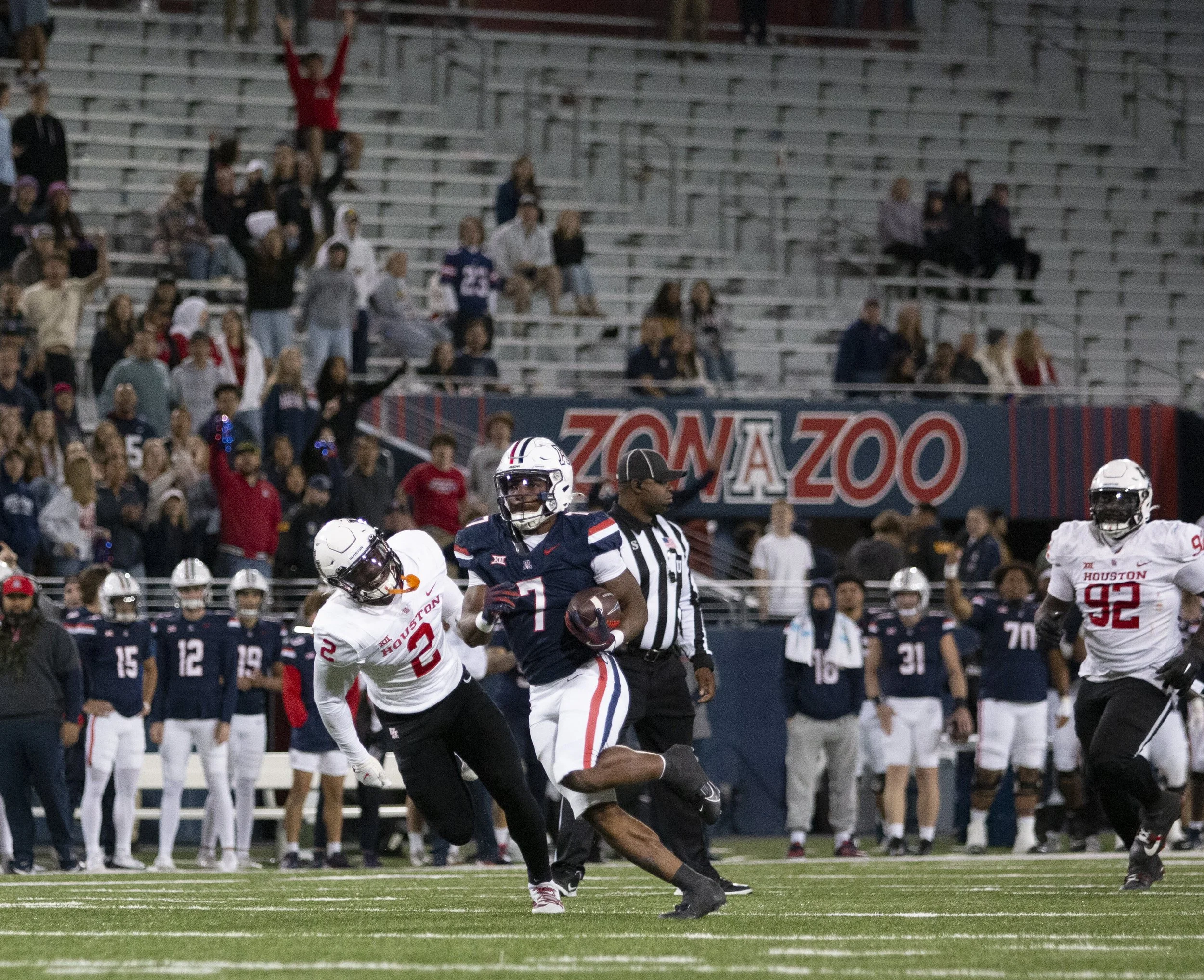 An American football game with players from Houston team in white and an opposing player in navy blue catching the football while a referee watches nearby, with spectators in the background.