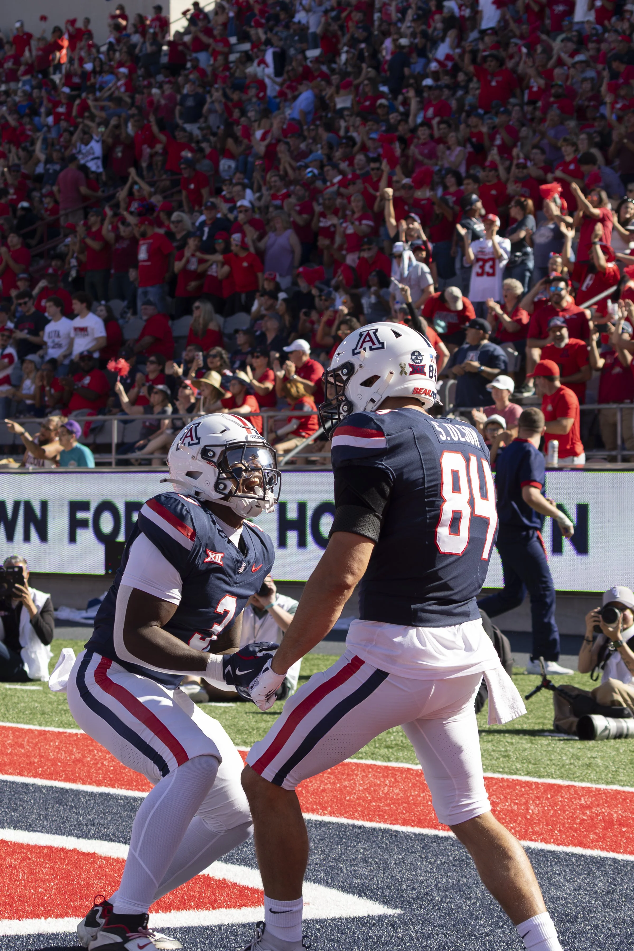 Two American football players in dark blue jerseys with red, white, and blue stripes celebrating after a play, with a large crowd of spectators in red shirts and hats in the background.