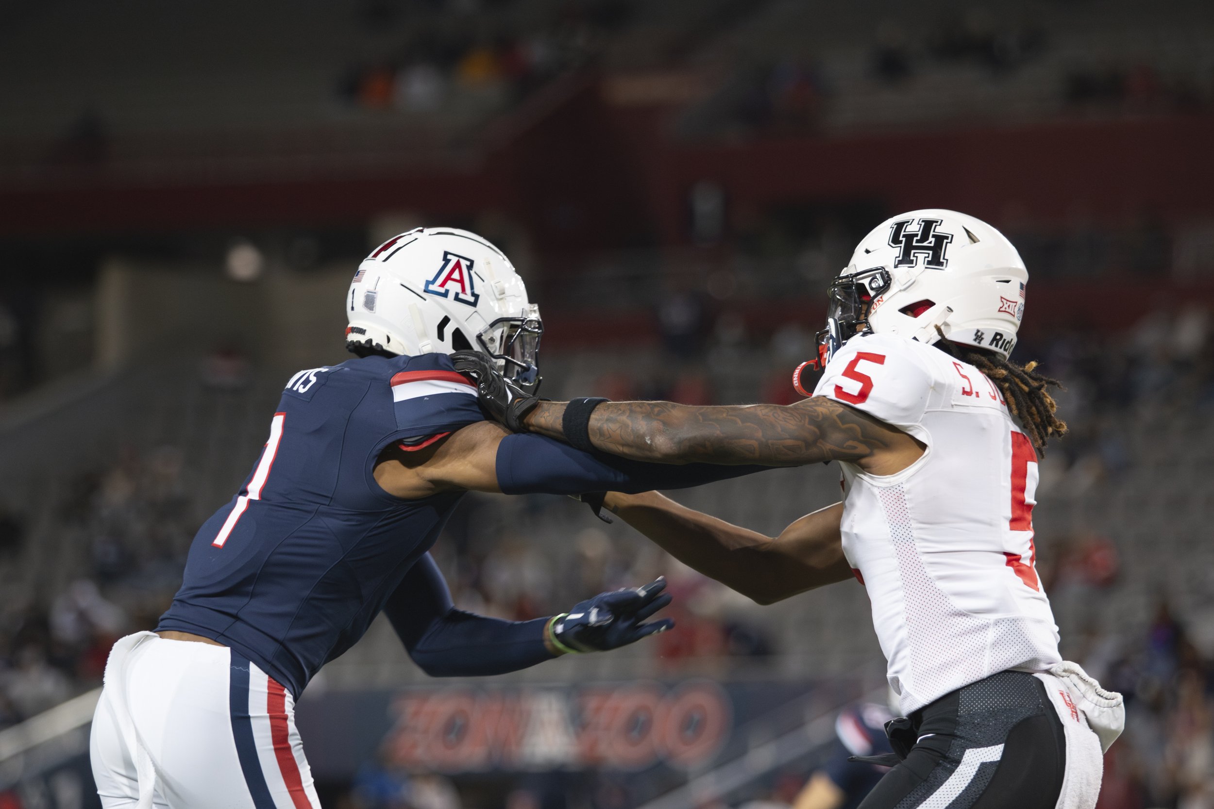 Two football players in helmets and uniforms engaged in a tackle during a game, with one player from Arizona in a blue jersey and another from Houston in a white jersey.