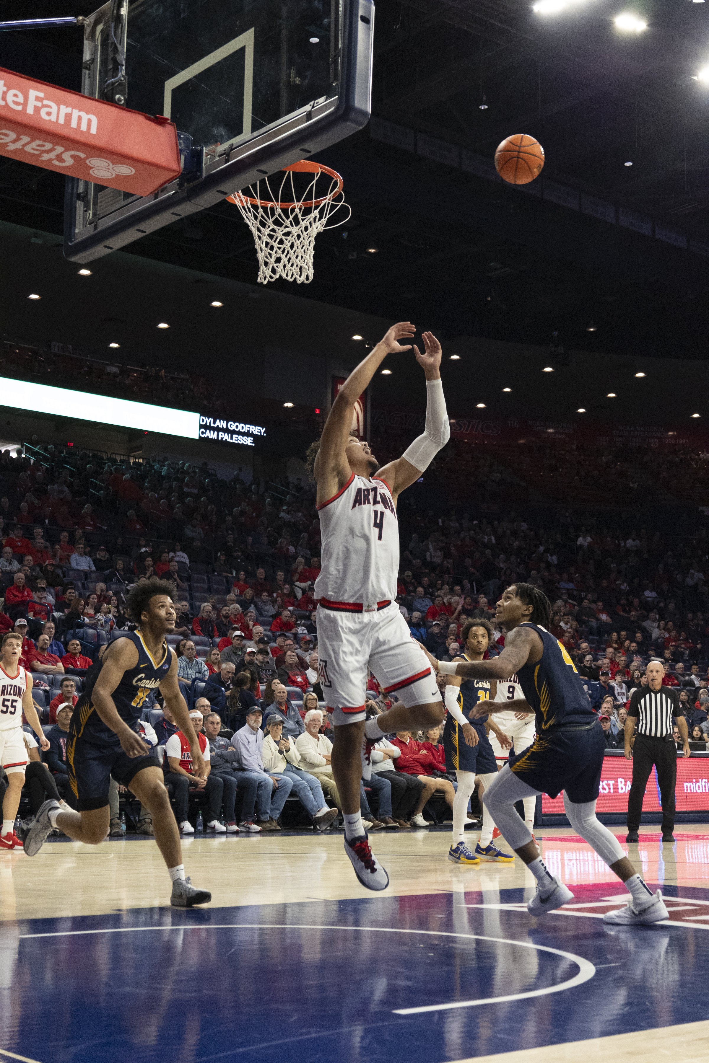 A basketball player from the University of Arizona in a white uniform wearing number 4 is jumping towards the hoop to make a shot during a game, with defenders and spectators in the background.