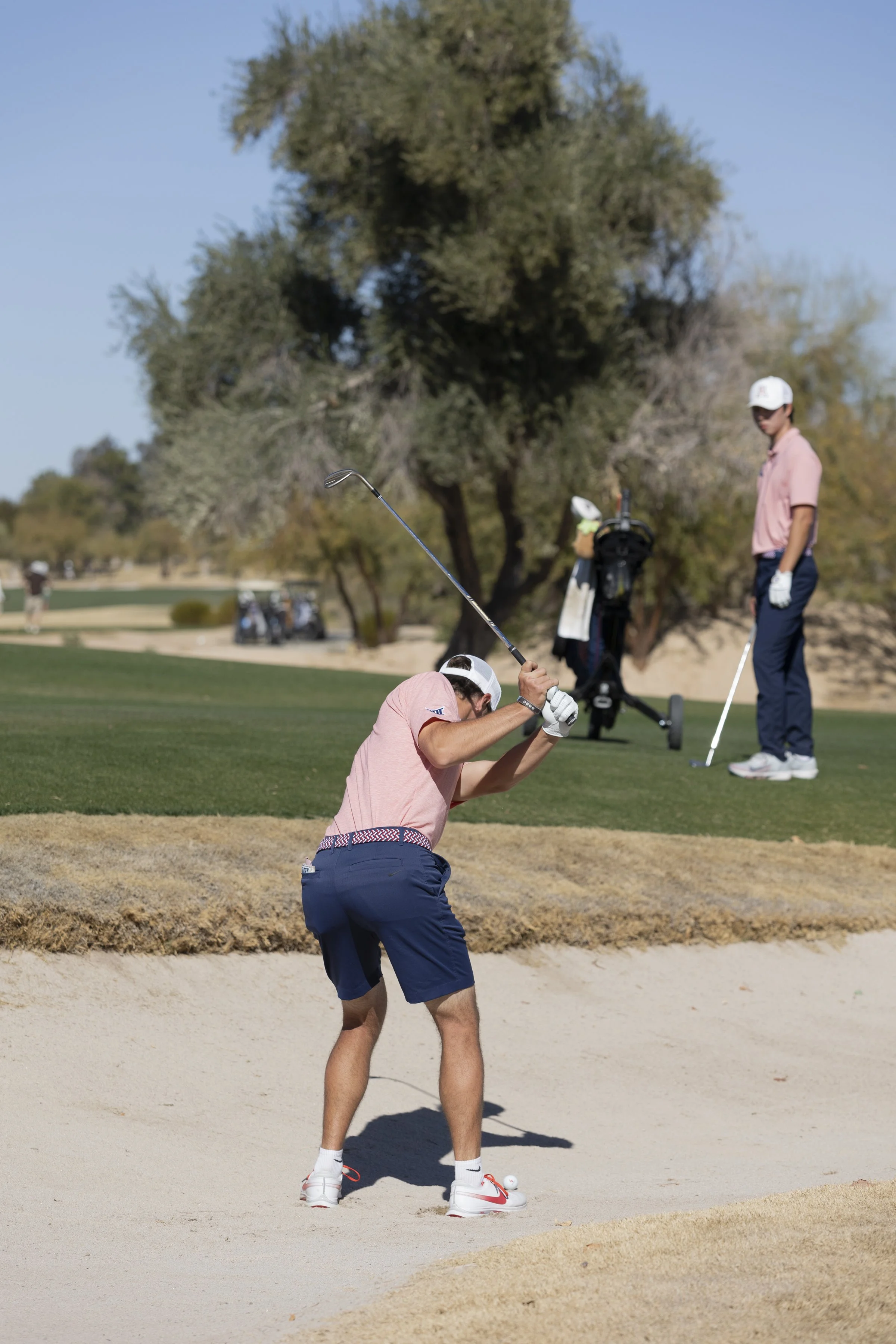 A golfer swinging a club in a sand bunker on a golf course with trees in the background.