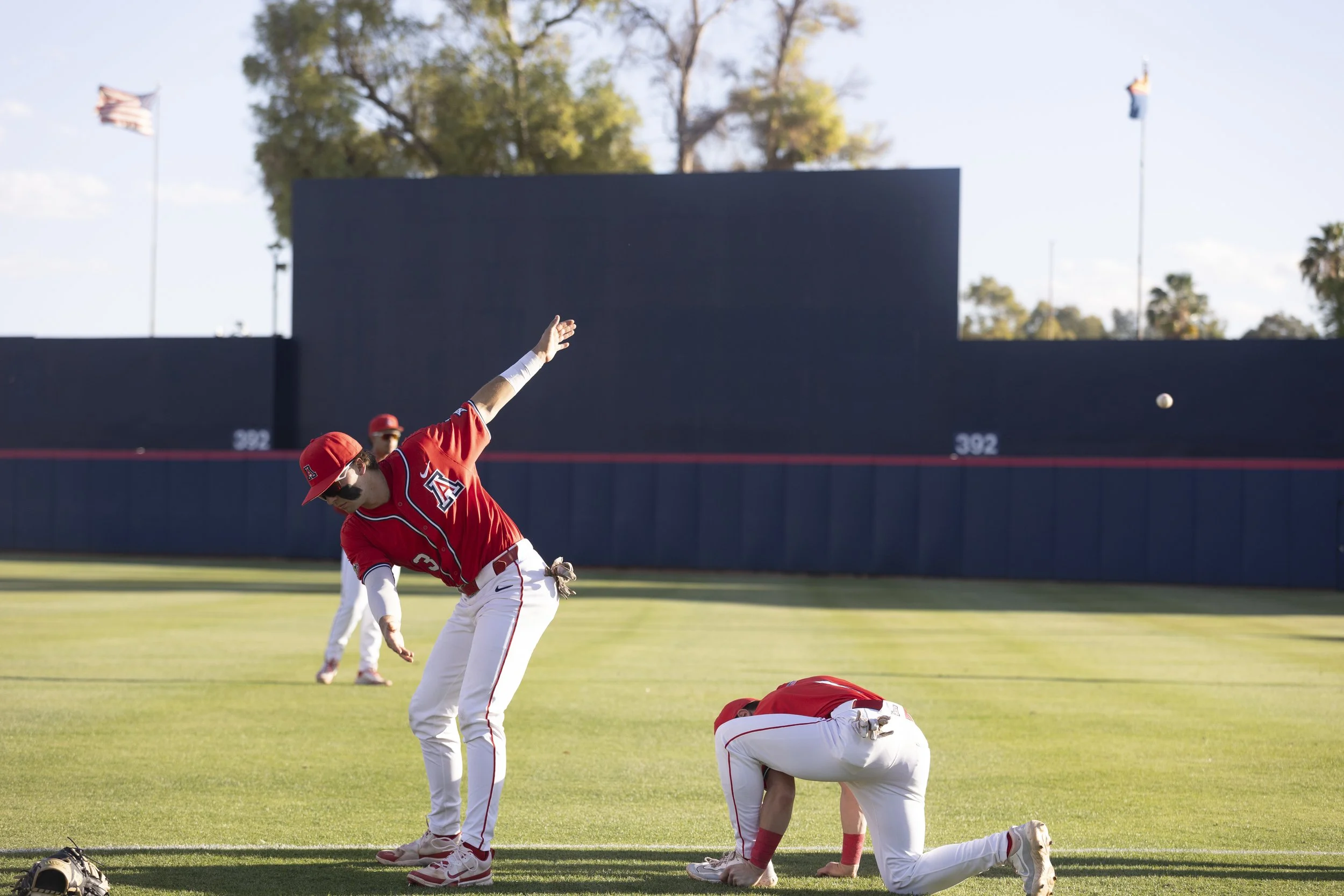 Two baseball players in red uniforms on the field practicing, with one in a crouch and the other stretching, while a ball is in mid-air in front of them.