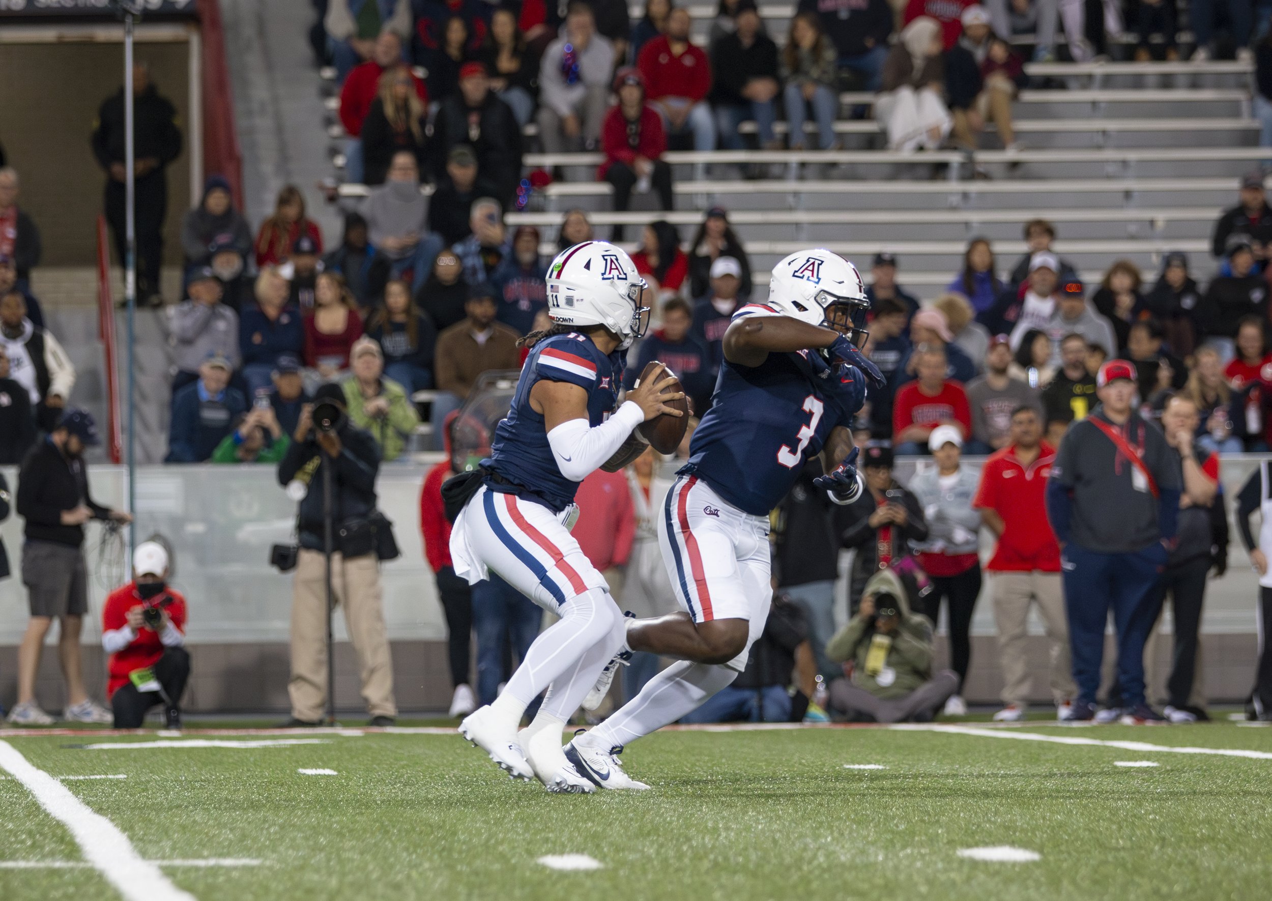 Two football players from the University of Arizona, in navy blue jerseys and white pants, during a game. One player is holding the football, while the other is running alongside him on the field. Spectators in the stands watch.
