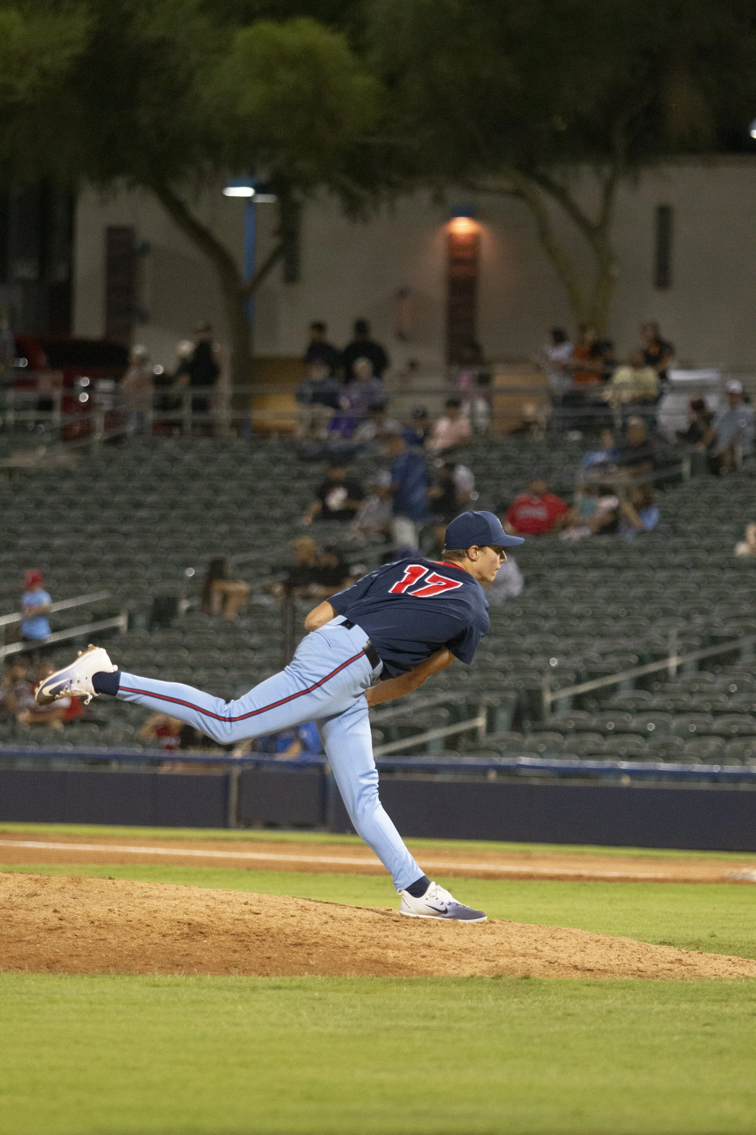 A young baseball pitcher in uniform throwing a pitch on the mound at a baseball field during evening or night, with spectators and trees in the background.
