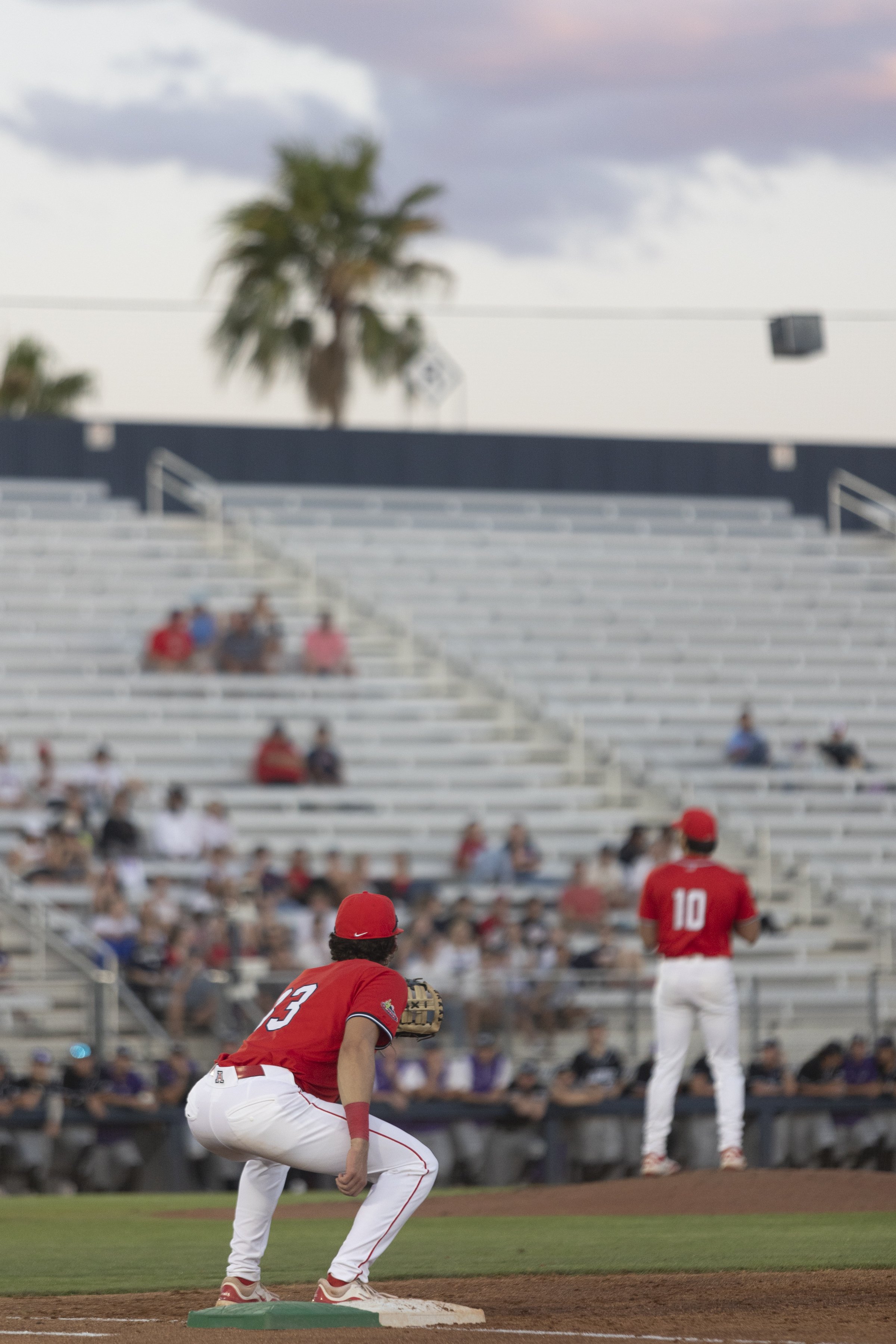 A baseball game in progress with players in red uniforms on the field and spectators in the stands, featuring a pitcher and a player near first base.