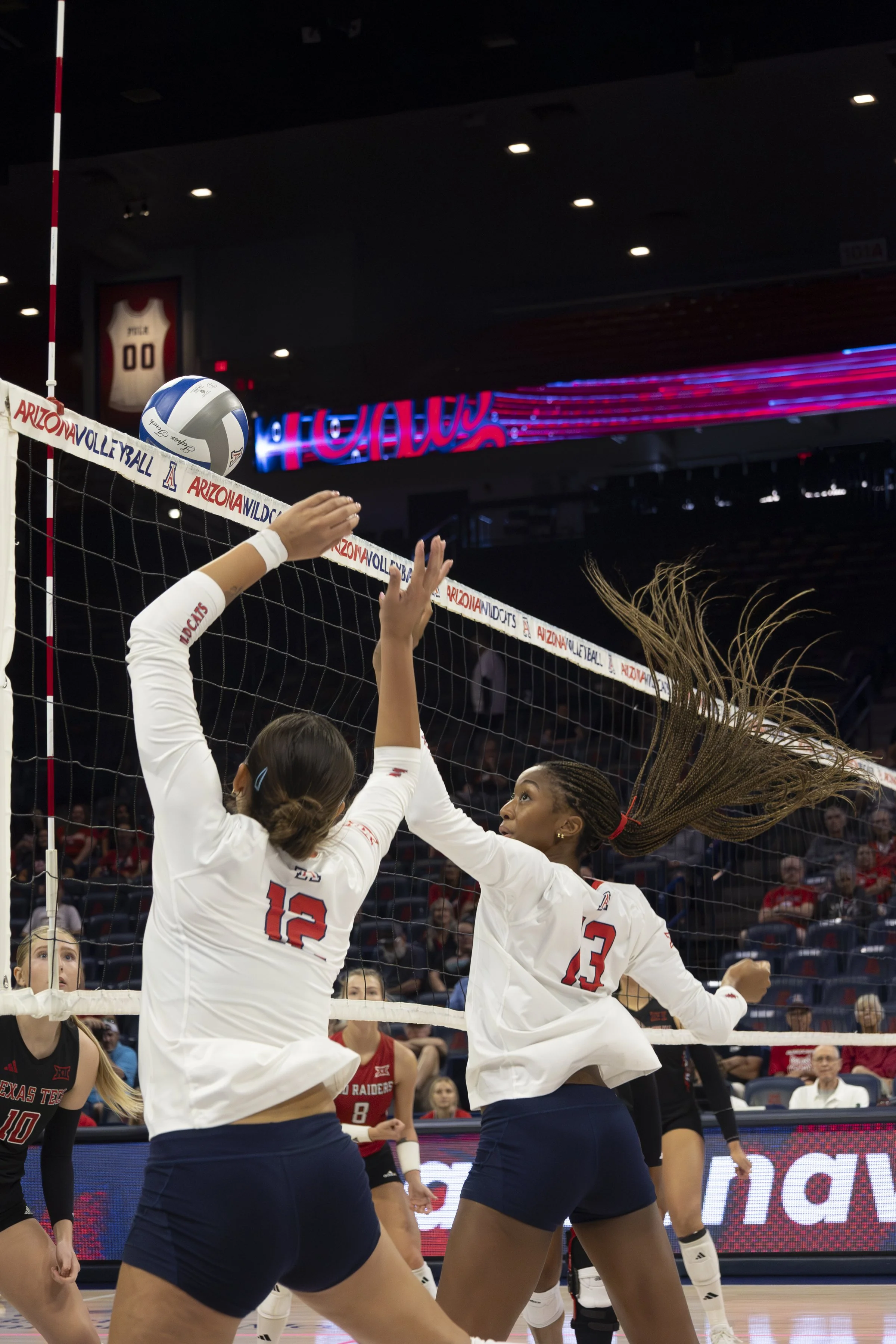 Two female volleyball players, one with jersey number 12 and the other with 23, jumping near the net to block a volleyball during a match at Arizona Wildcats arena with spectators in the background.