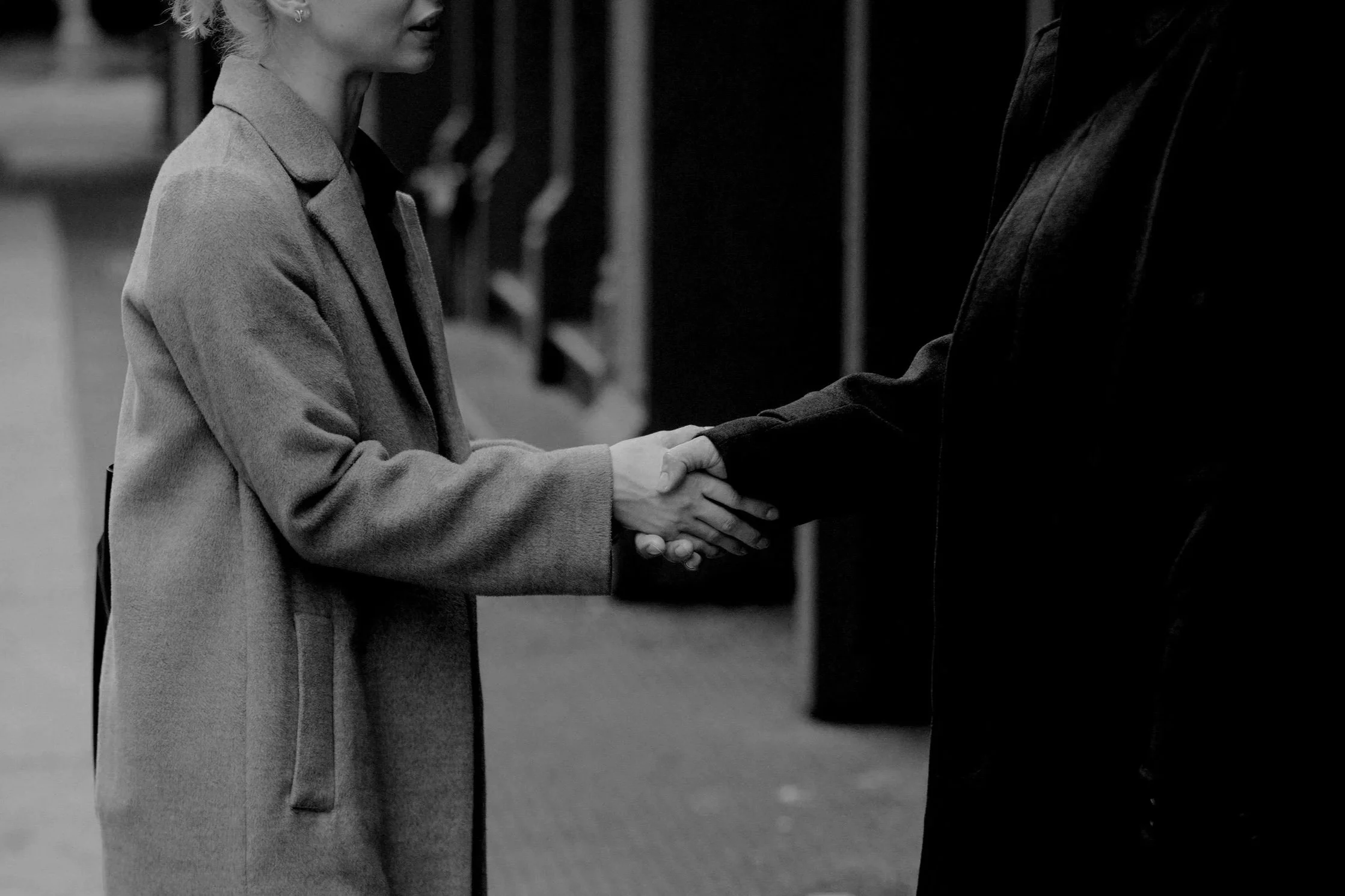 A woman and a man shaking hands indoors, with the woman wearing a coat and the man dressed in a suit.