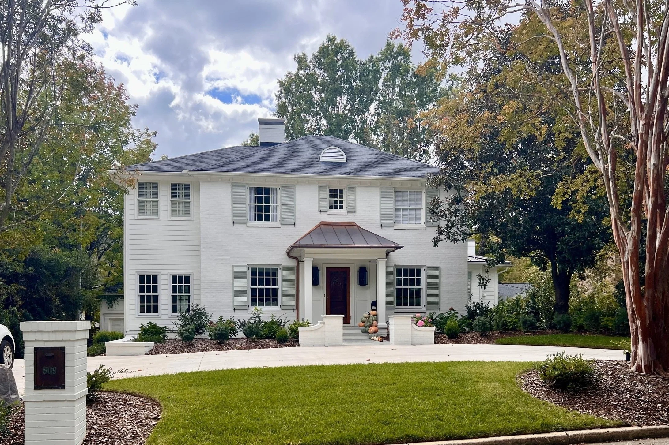 White two-story house with gray shutters, front porch with columns, a curved walkway, lush green lawn, and large trees around
