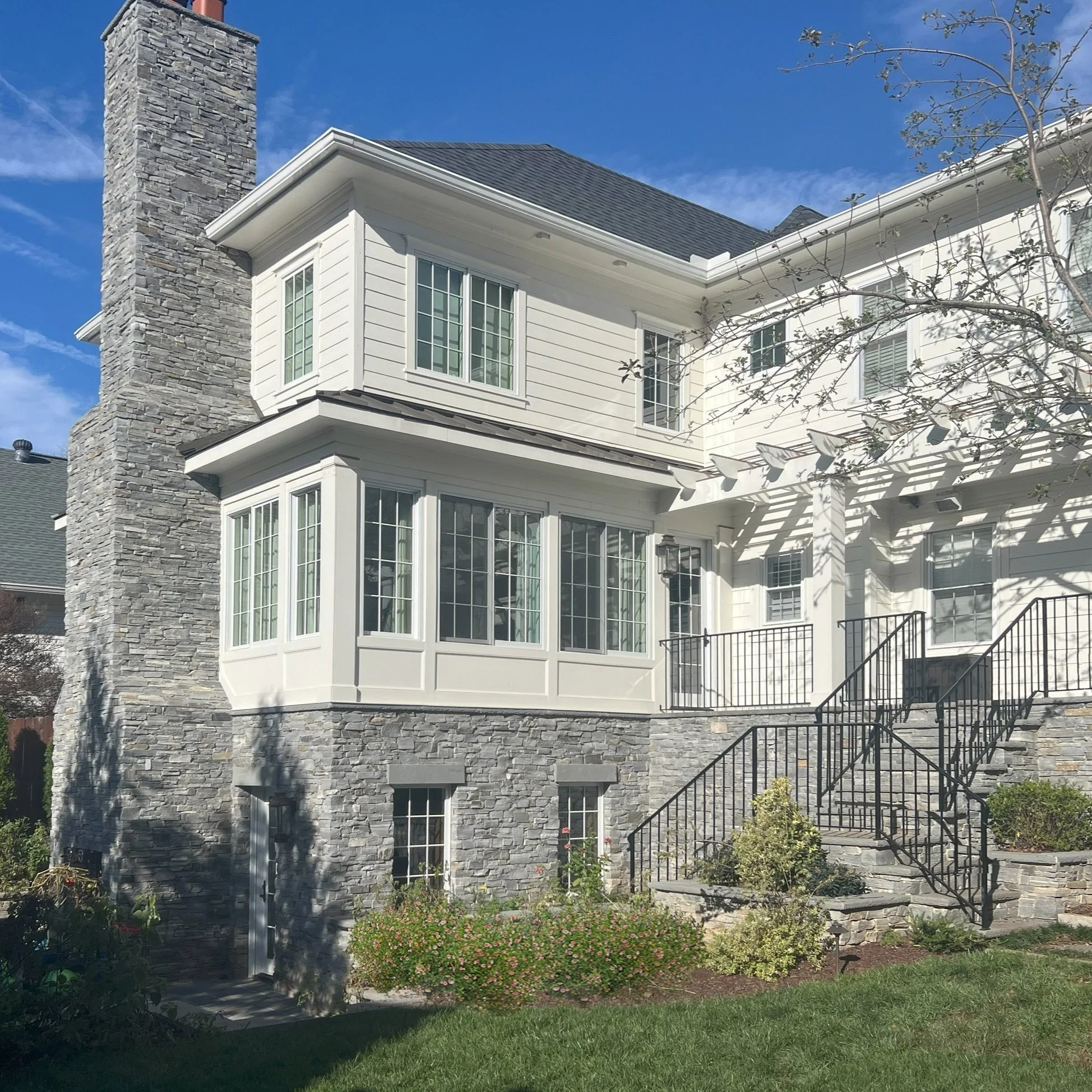 A multi-story house with gray stone foundation and white siding, featuring large bay windows, a stone chimney, a staircase with black metal railings, and a garden with green plants and flowers.