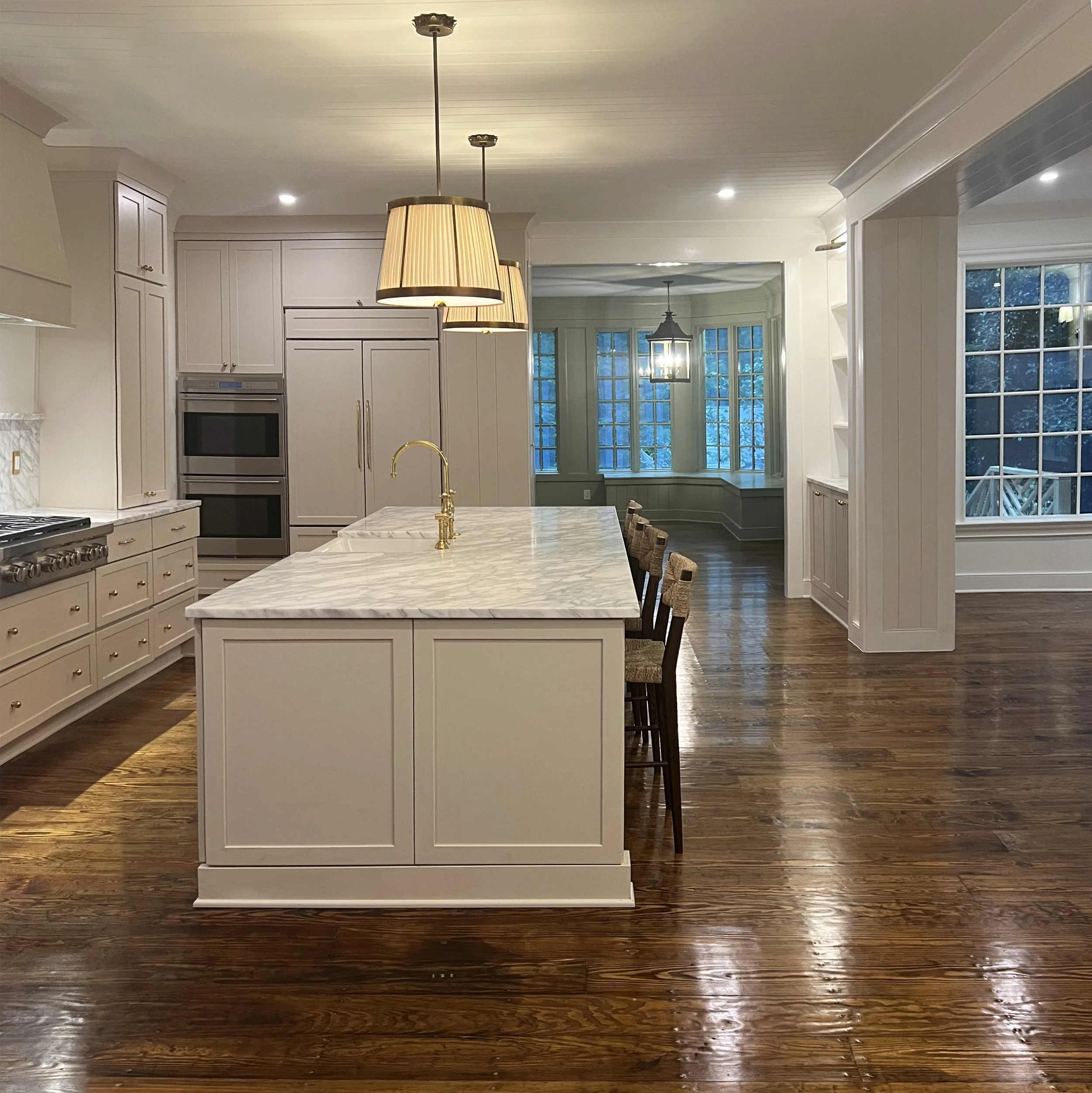 Modern kitchen with white cabinetry, marble island, wooden floor, and large windows showing an outdoor view at dusk.