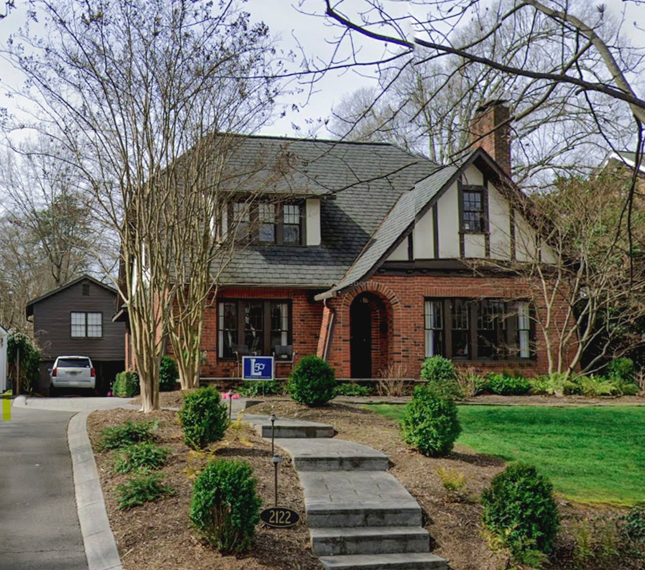 A two-story brick house with a gabled roof, surrounded by landscaped greenery and a walkway leading to the front door.
