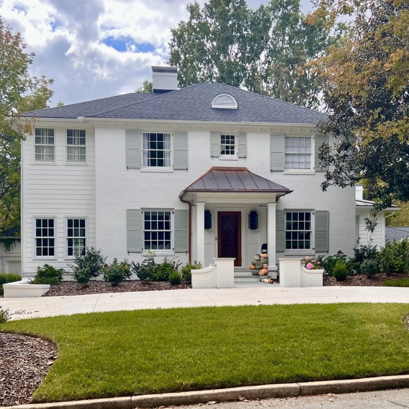 A two-story white house with gray shutters, a gray shingle roof, and a small porch with a brown metal roof. The house is surrounded by greenery and a well-maintained lawn.