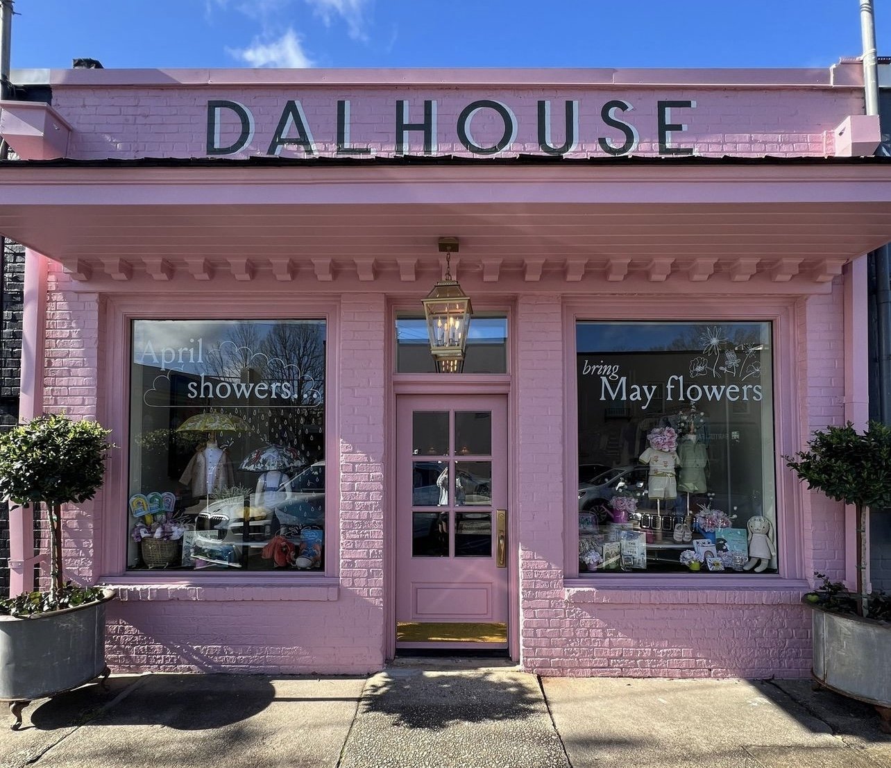A pink storefront with large windows and a door in the center. The sign on top reads 'DALHOUSE.' The left window displays a sign that says 'April showers,' and the right window has a sign that says 'bring May flowers.' The window displays include clothing, umbrellas, flowers, and stuffed animals. There are potted plants on either side of the entrance and a hanging lantern above the door.