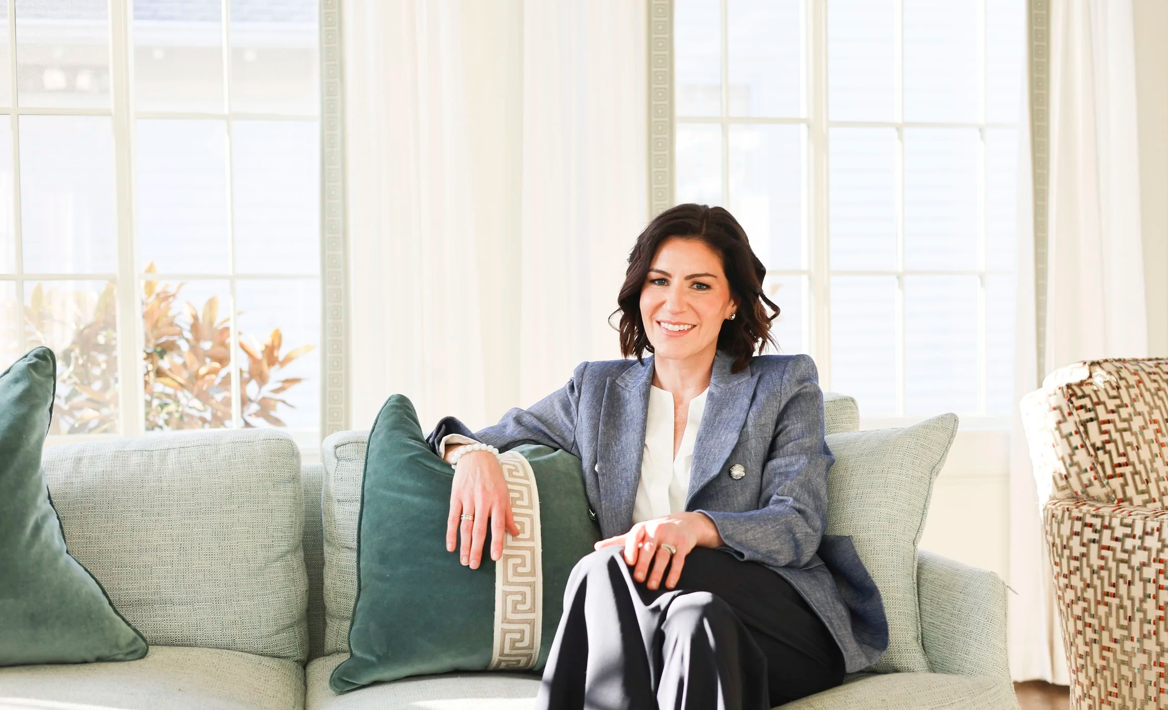 A woman sitting on a light-colored sofa in a sunlit room, smiling. She has dark, wavy hair and is wearing a gray blazer over a white top, black and white striped pants. The room has large windows with white curtains, and there are green and patterned pillows on the sofa.