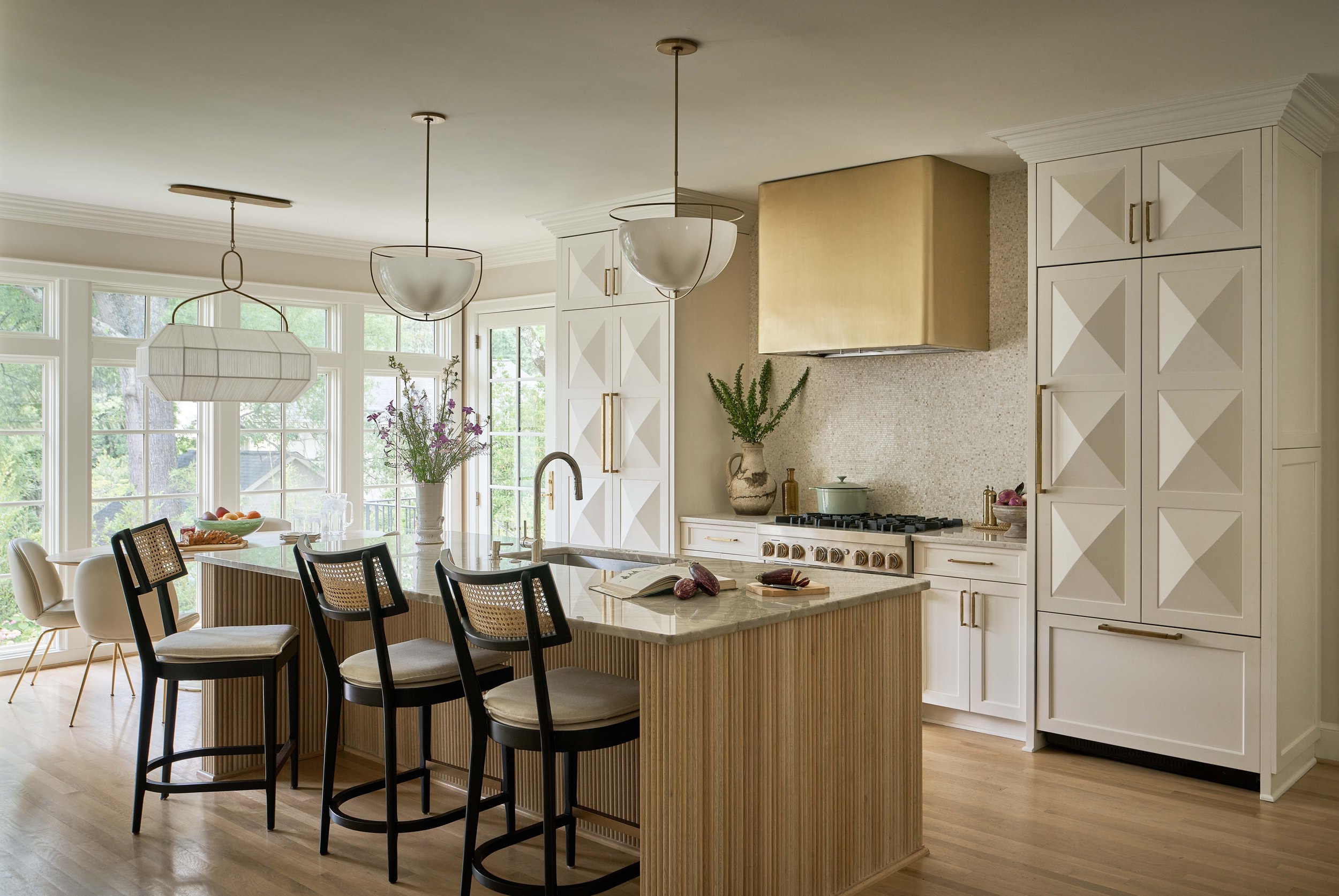 Bright, modern kitchen with a central island featuring a marble countertop and black chairs, large windows in the background, white cabinets with diagonal paneling, and gold accents, decorated with plants and fresh produce.