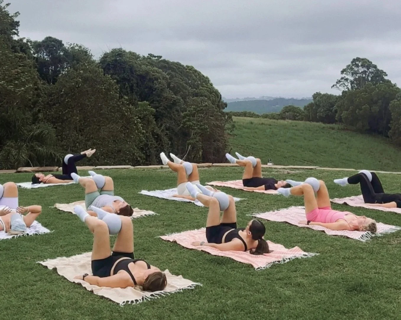 A group of women participating in an outdoor fitness class, lying on mats and performing leg lifts on a grassy field with trees and cloudy sky in the background.