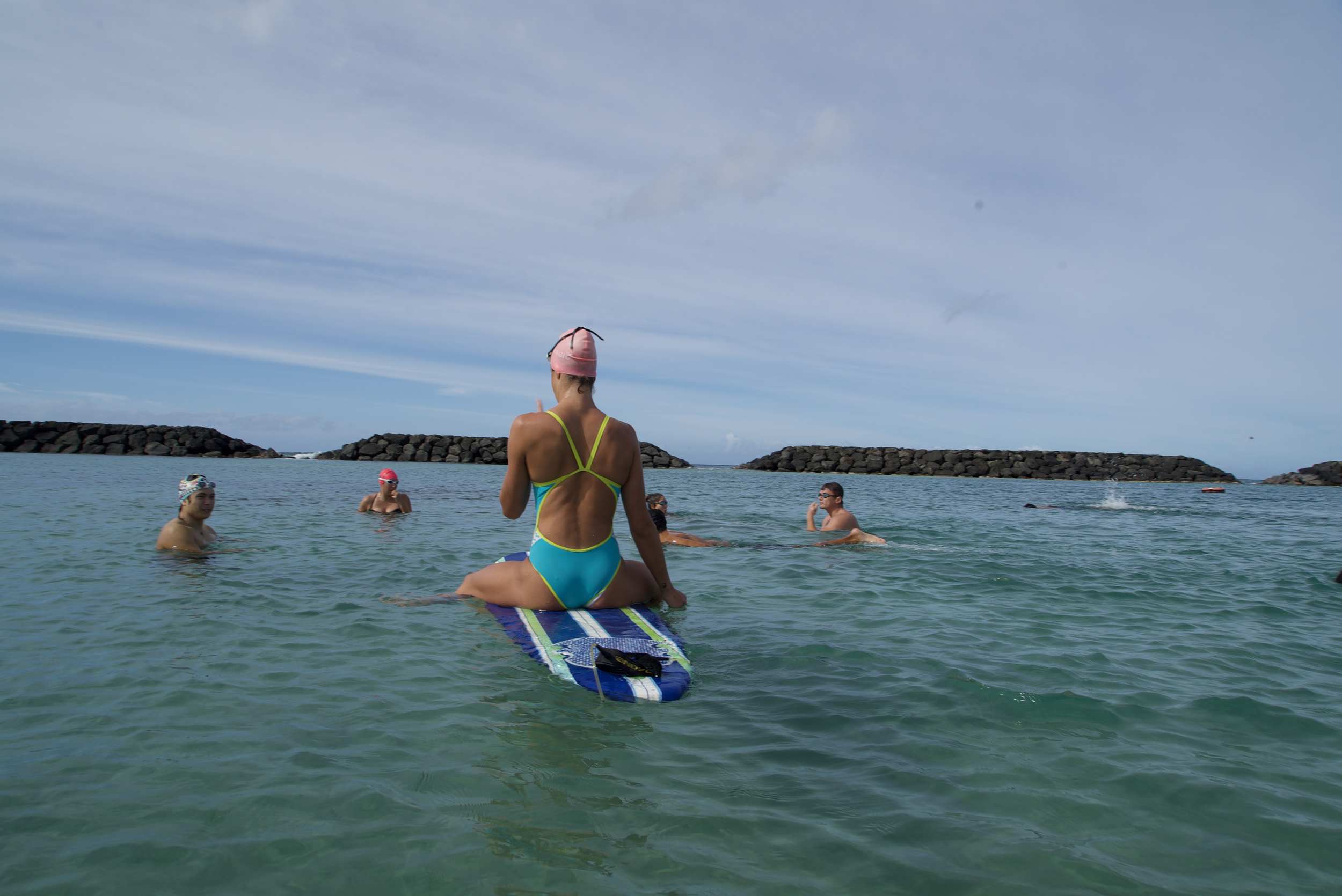 A woman in a green and blue swimsuit sitting on a surfboard in the ocean, surrounded by swimmers, with rocky breakwaters in the background under a clear blue sky.
