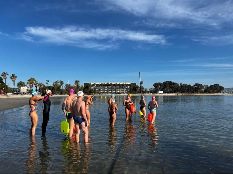 Group of people in swimsuits and swim caps standing in shallow water at the beach, some holding swim buoys, with a sandy shoreline, palm trees, and a building in the background under a blue sky.