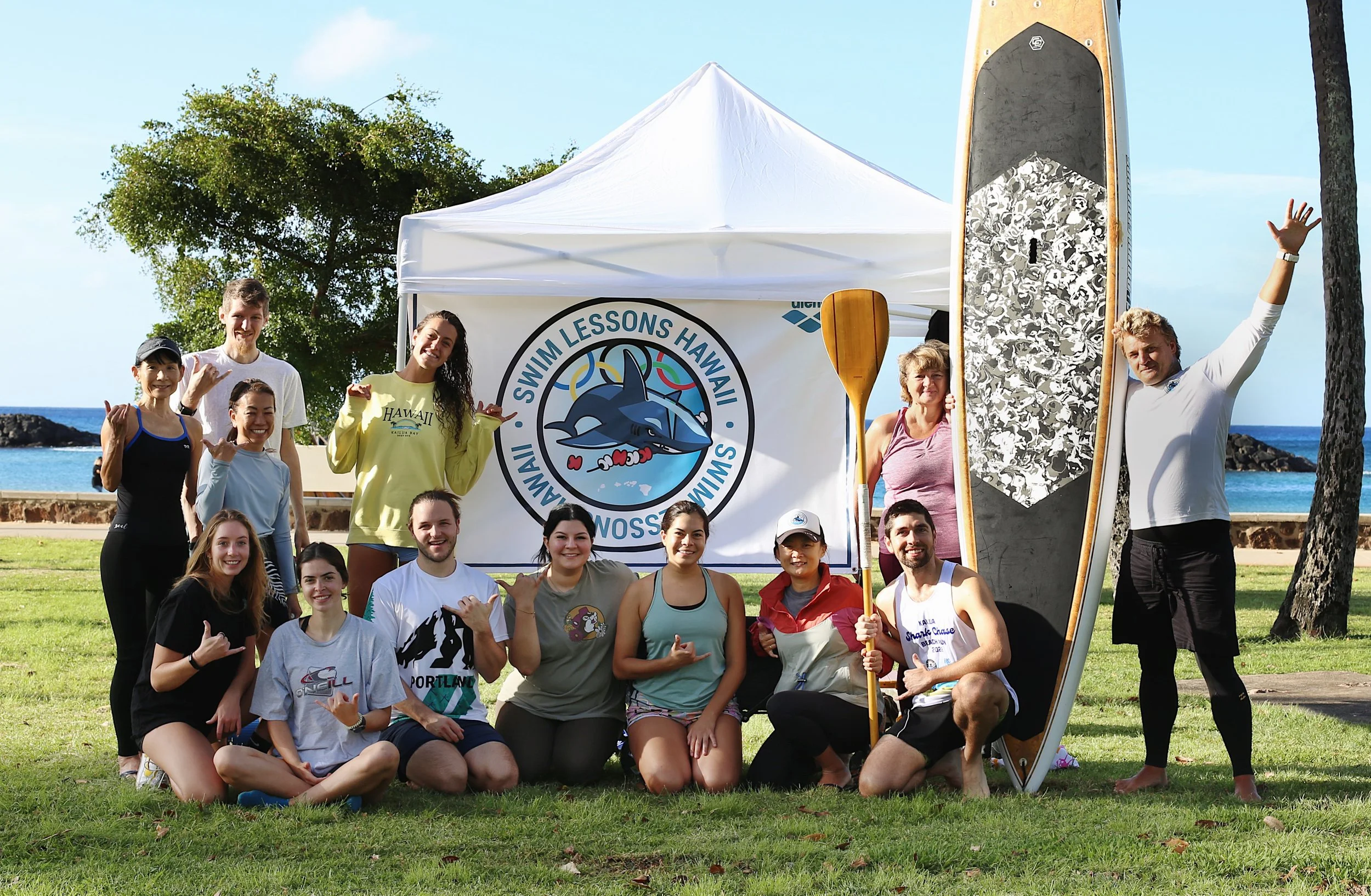 Group of people posing outdoors at a beachfront park with a white canopy tent and a large paddleboard. The banner behind them reads 'Swim Lessons Hawaii' with a dolphin logo. The group is smiling and making hand gestures, with some holding paddles an