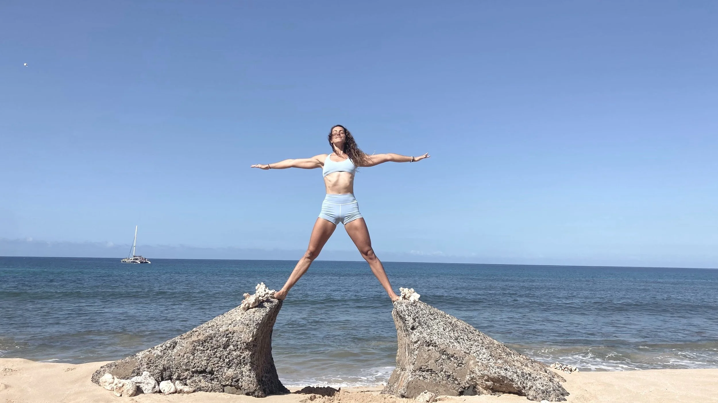 Woman standing with arms outstretched on two large rocks at the beach, facing the ocean with a boat in the background.