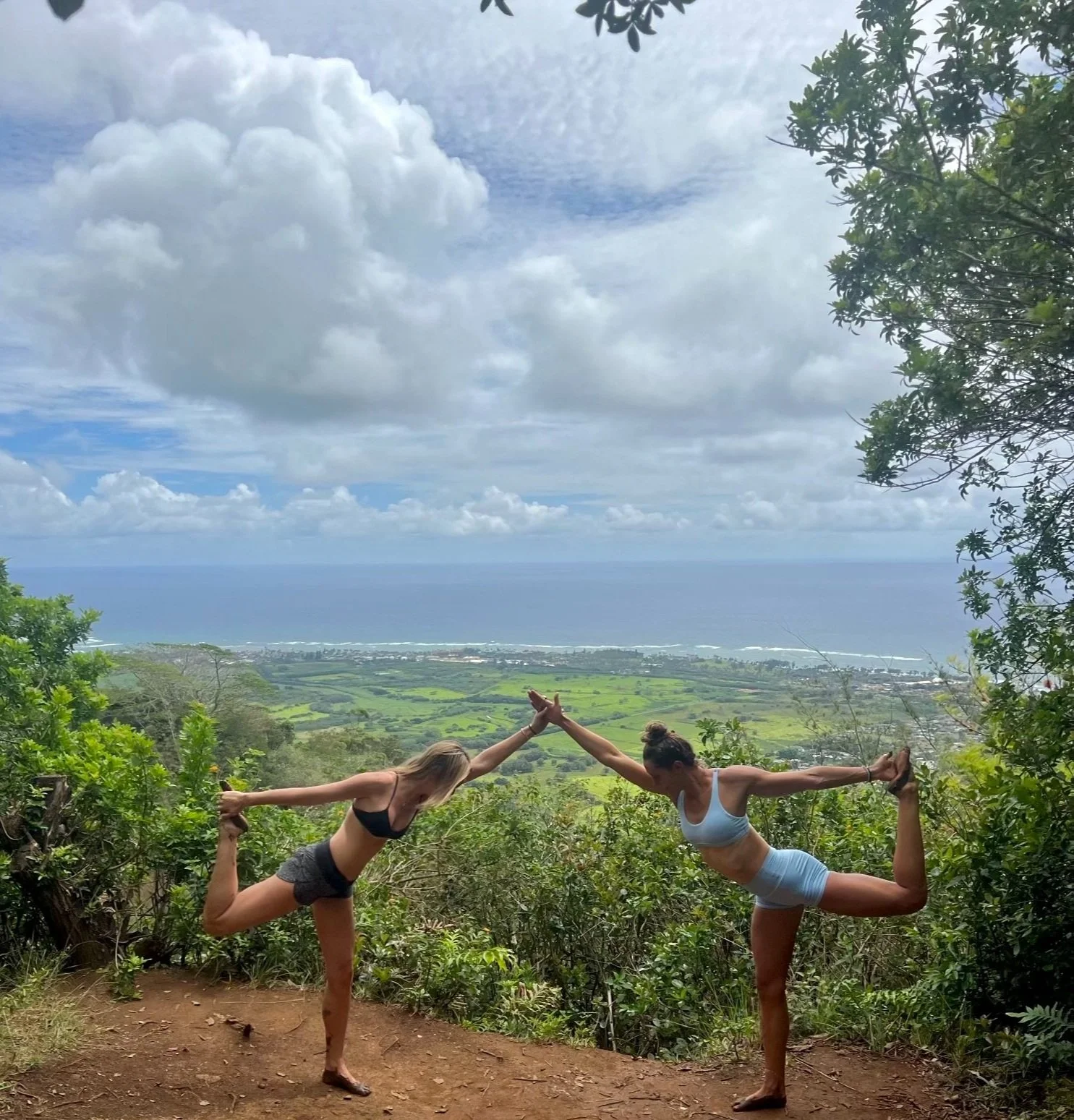 Two women practicing yoga on a hillside overlooking a lush green landscape and the ocean, with cloudy skies above.